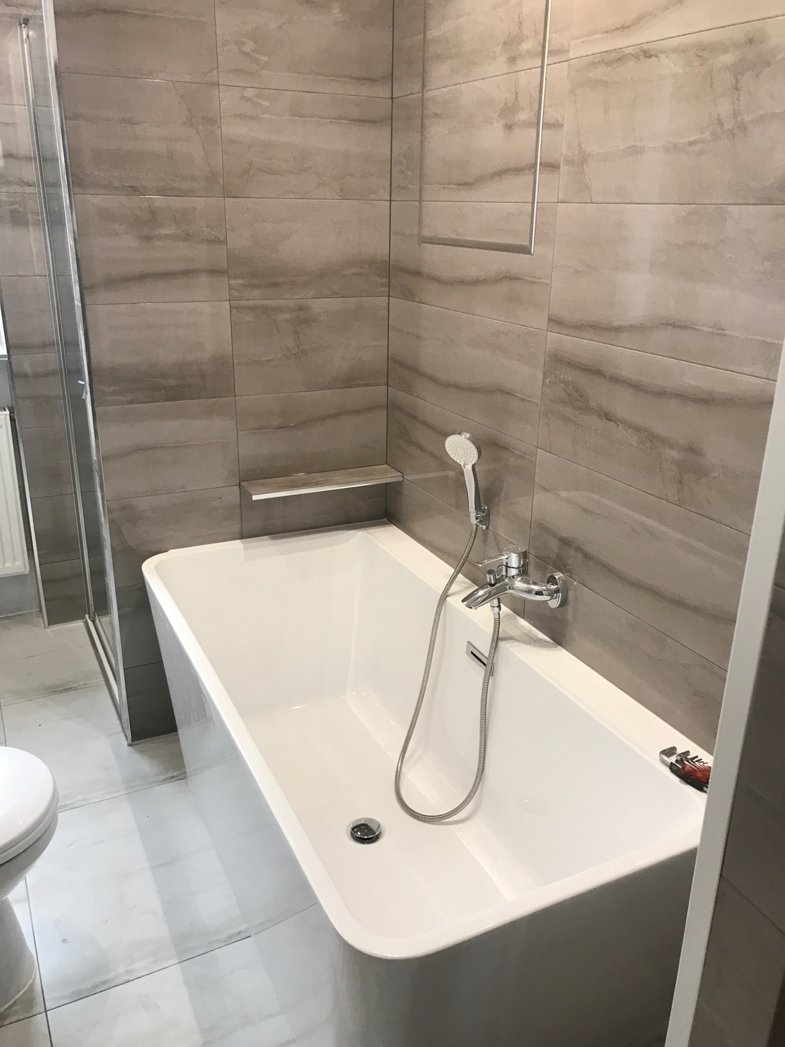 Nowoczesna łazienka z wanną prostokątną, baterią wannową z słuchawką prysznicową, płytkami imitującymi kamień w odcieniach szarości oraz kabiną prysznicową typu walk-in. Na krawędzi wanny leżą...