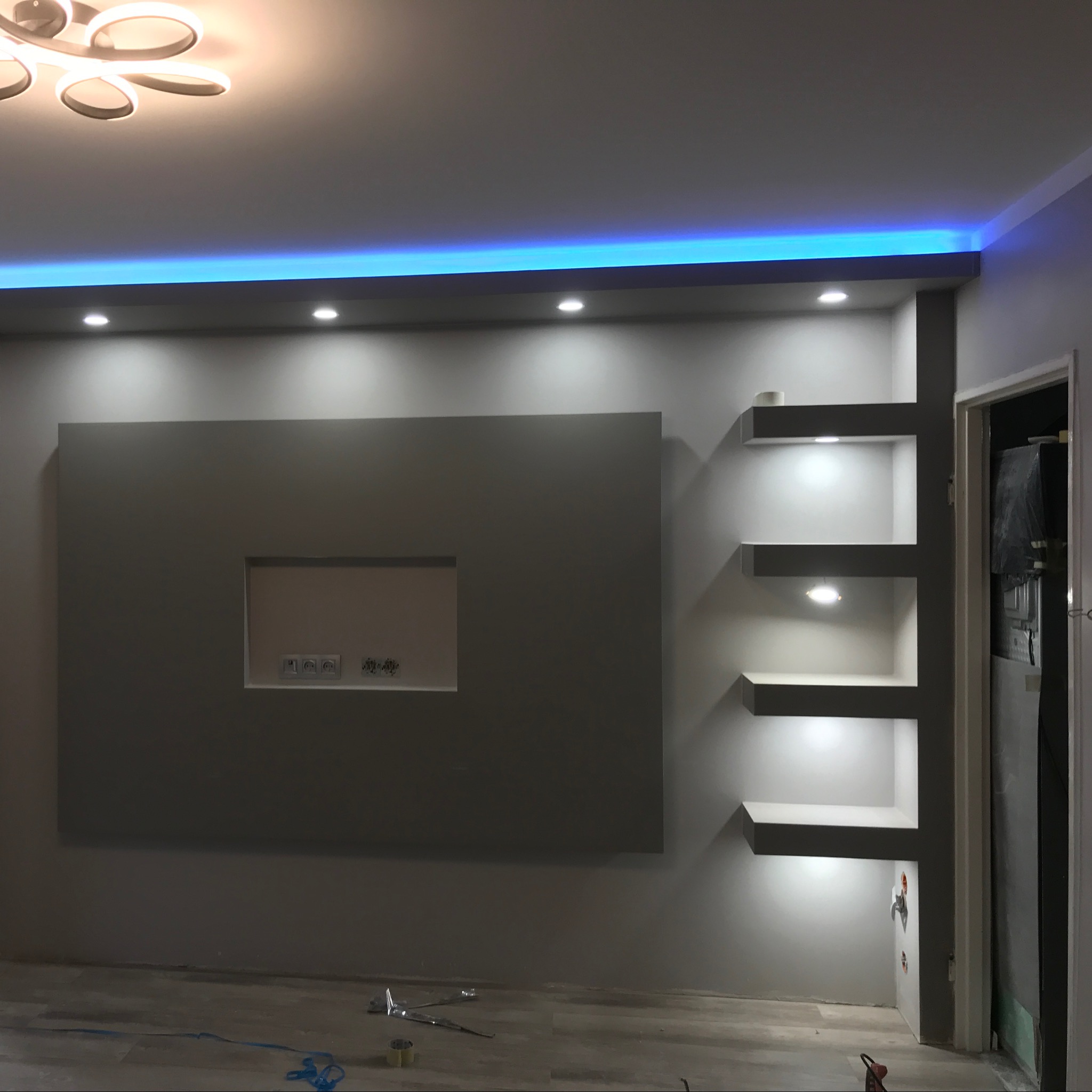 Szara ściana z wnęką na telewizor i półkami podświetlonymi punktowymi lampami LED, niebieskie oświetlenie LED podsufitowe, widoczne gniazdka elektryczne i fragment podłogi z narzędziami.