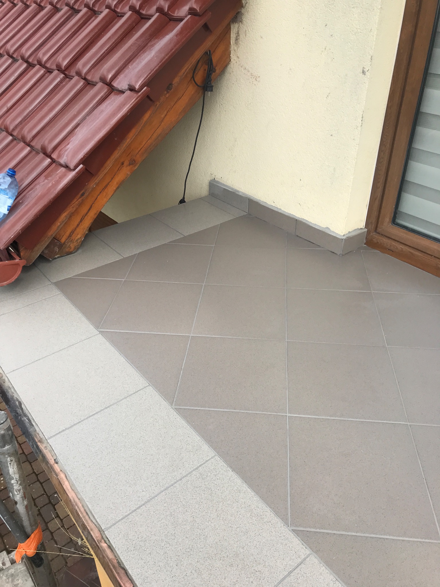 Wyłożony płytkami taras z widocznym fragmentem dachu z czerwonej dachówki i oknem z brązową ramą, z zastosowaniem dwóch odcieni płytek.