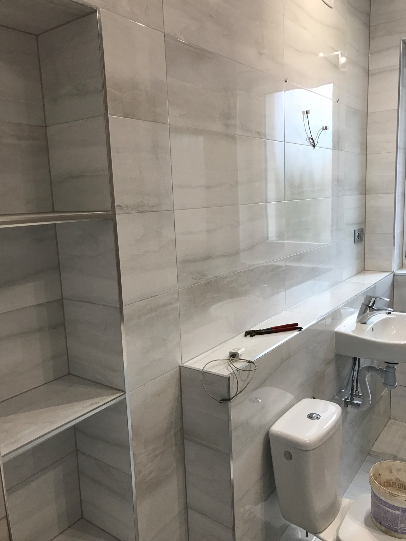 Nowoczesna łazienka w trakcie wykańczania, widoczne białe płytki imitujące marmur, wisząca umywalka, toaleta, półki w zabudowie, narzędzia i kable na blacie.