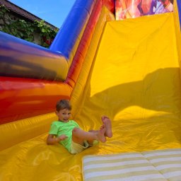 Zabawa u Adasia - Uśmiechnięty chłopiec w zielonej koszulce zjeżdża po żółtej, dmuchanej zjeżdżalni z motywem Sonic'a, widoczne stopy dziecka i fragment poduszki na końcu zjeżdżalni.