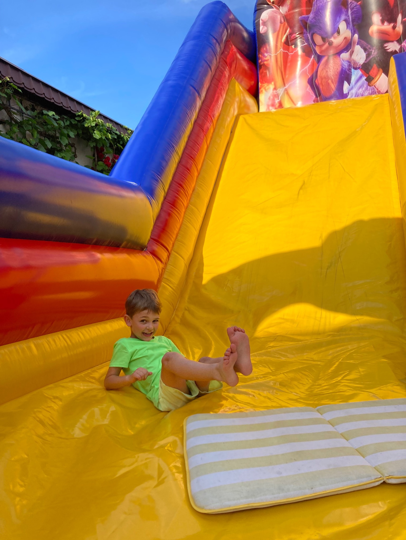Uśmiechnięty chłopiec w zielonej koszulce zjeżdża po żółtej, dmuchanej zjeżdżalni z motywem Sonic'a, widoczne stopy dziecka i fragment poduszki na końcu zjeżdżalni.