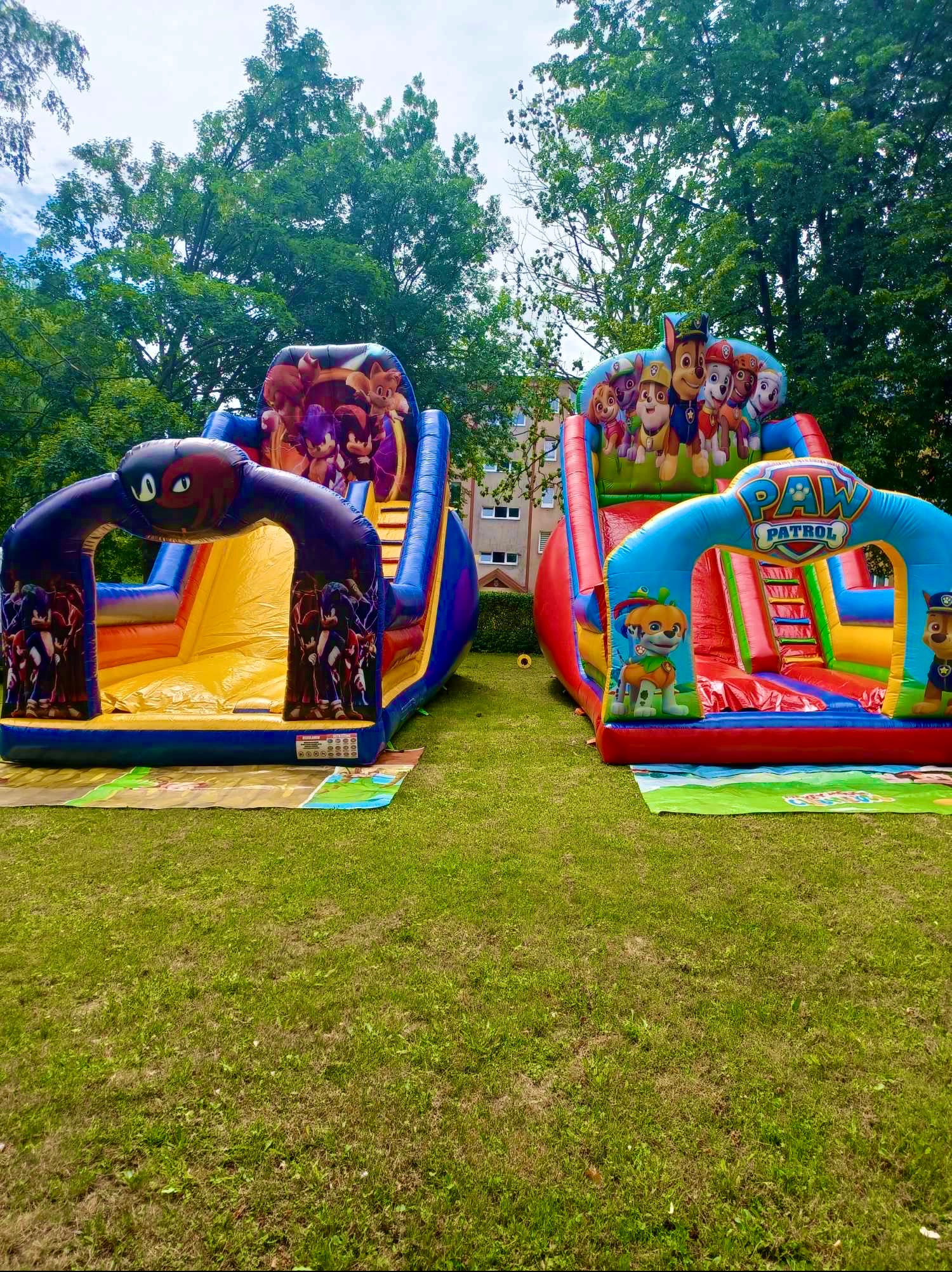 Dwa kolorowe, nadmuchiwane zamki do skakania z motywami z bajek (Sonic i Psi Patrol) ustawione na trawniku, z widocznymi drzewami w tle.