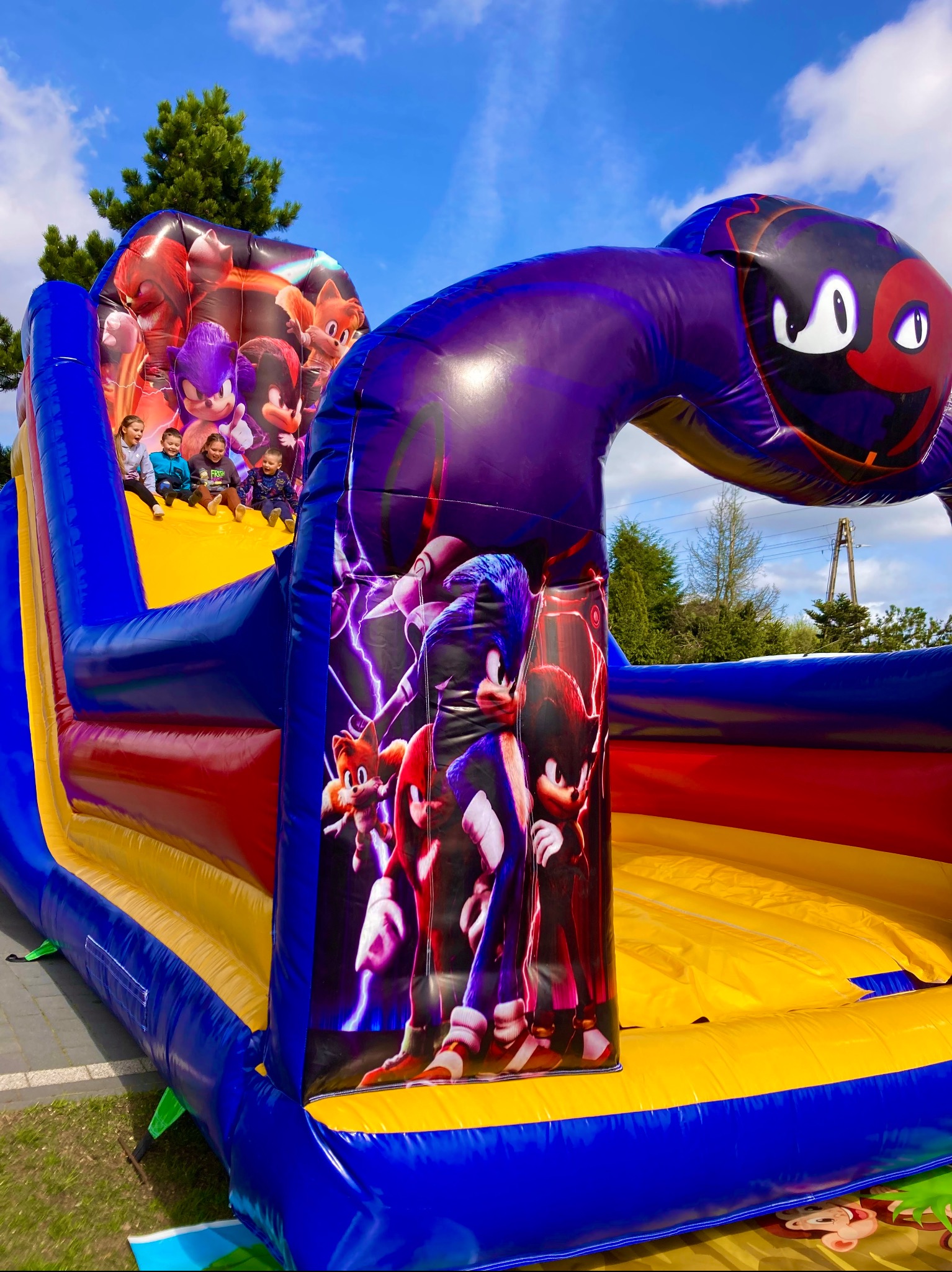 Kolorowy dmuchany zamek z motywem Sonic the Hedgehog, na którym bawi się czwórka dzieci, widok z dołu, błękitne niebo w tle.