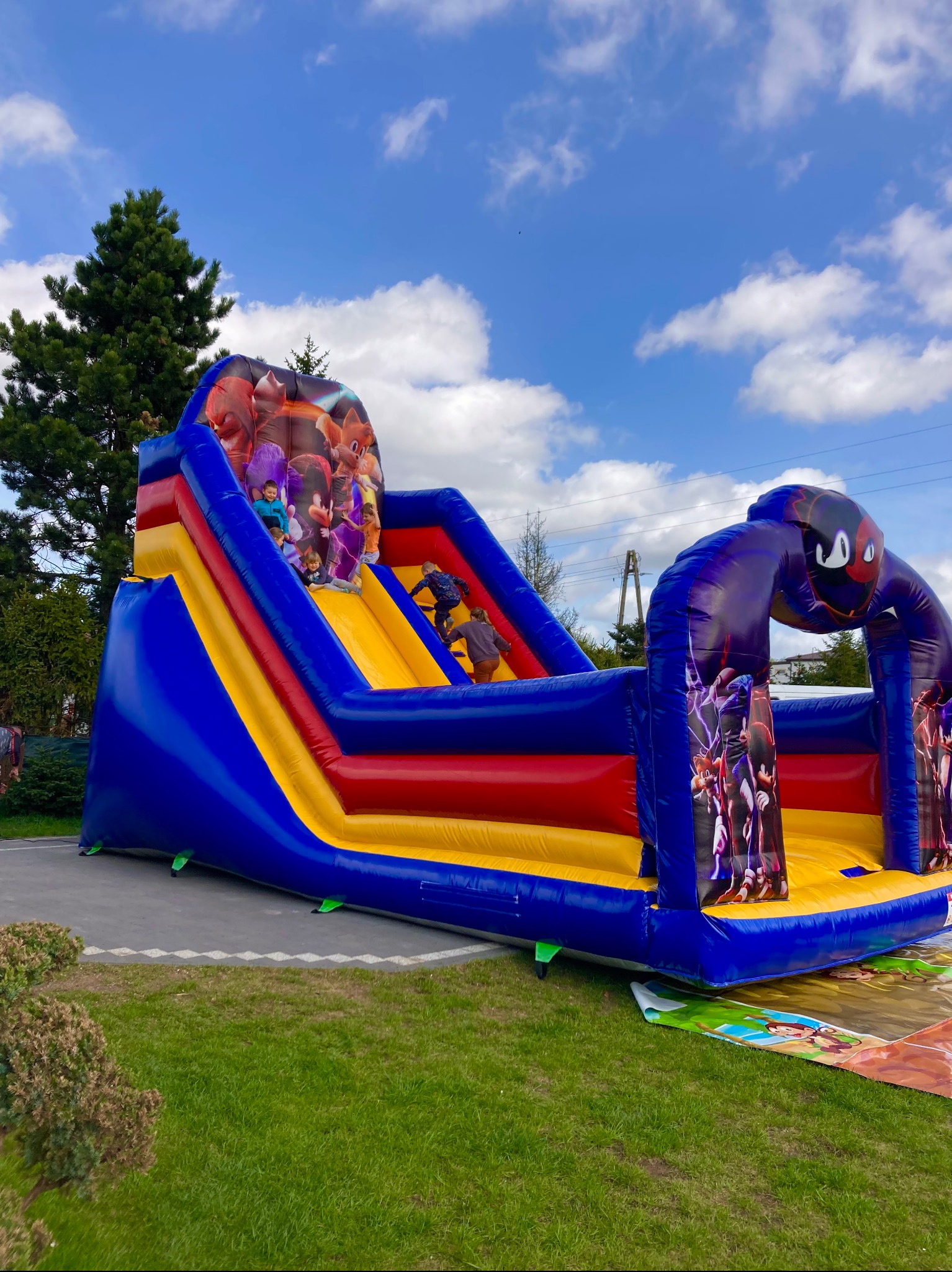 Kolorowy, dmuchany zamek ze zjeżdżalnią z motywami bajkowymi, dzieci bawiące się w słoneczny dzień na trawniku.