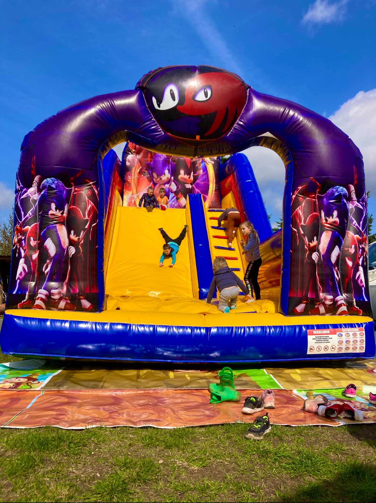 Dzieci bawią się na dmuchanym zamku ze zjeżdżalnią z motywem Sonic the Hedgehog; jedno dziecko zjeżdża głową w dół, inne wspinają się po schodach, a na dole leżą porozrzucane buty.