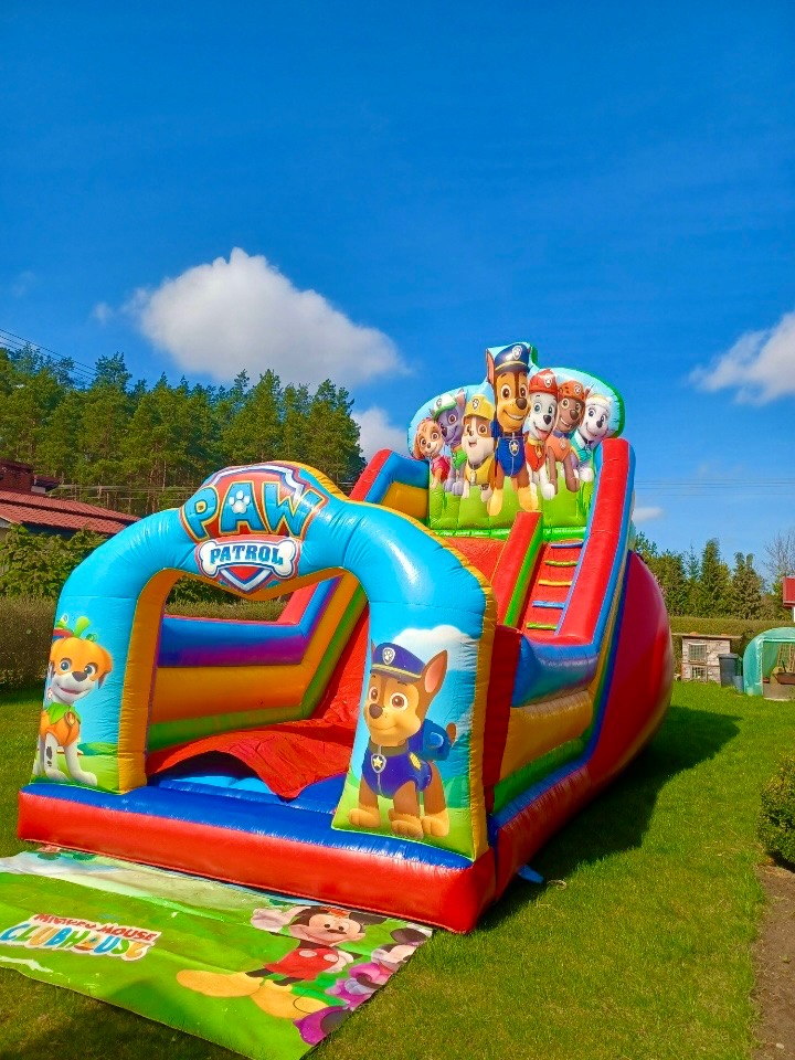 Kolorowa, dmuchana zjeżdżalnia z motywem 'Psi Patrol' na zielonej trawie, błękitne niebo z chmurami w tle. Na dole rozłożona mata z motywem Myszki Miki.