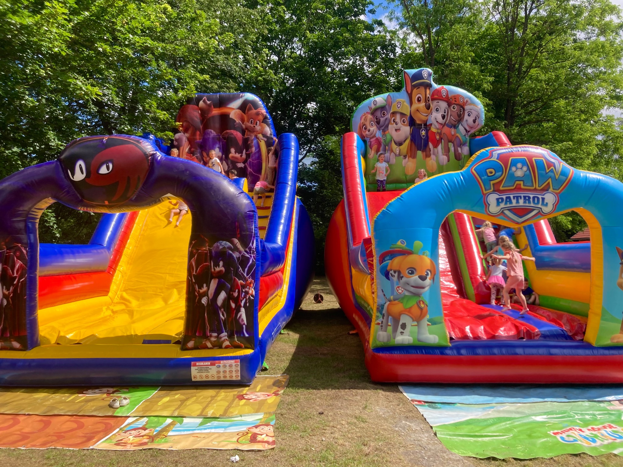 Dwa kolorowe dmuchane zamki ze zjeżdżalniami dla dzieci, jeden z motywem Sonic, drugi z motywem Paw Patrol, ustawione obok siebie na trawie, z bawiącymi się dziećmi.