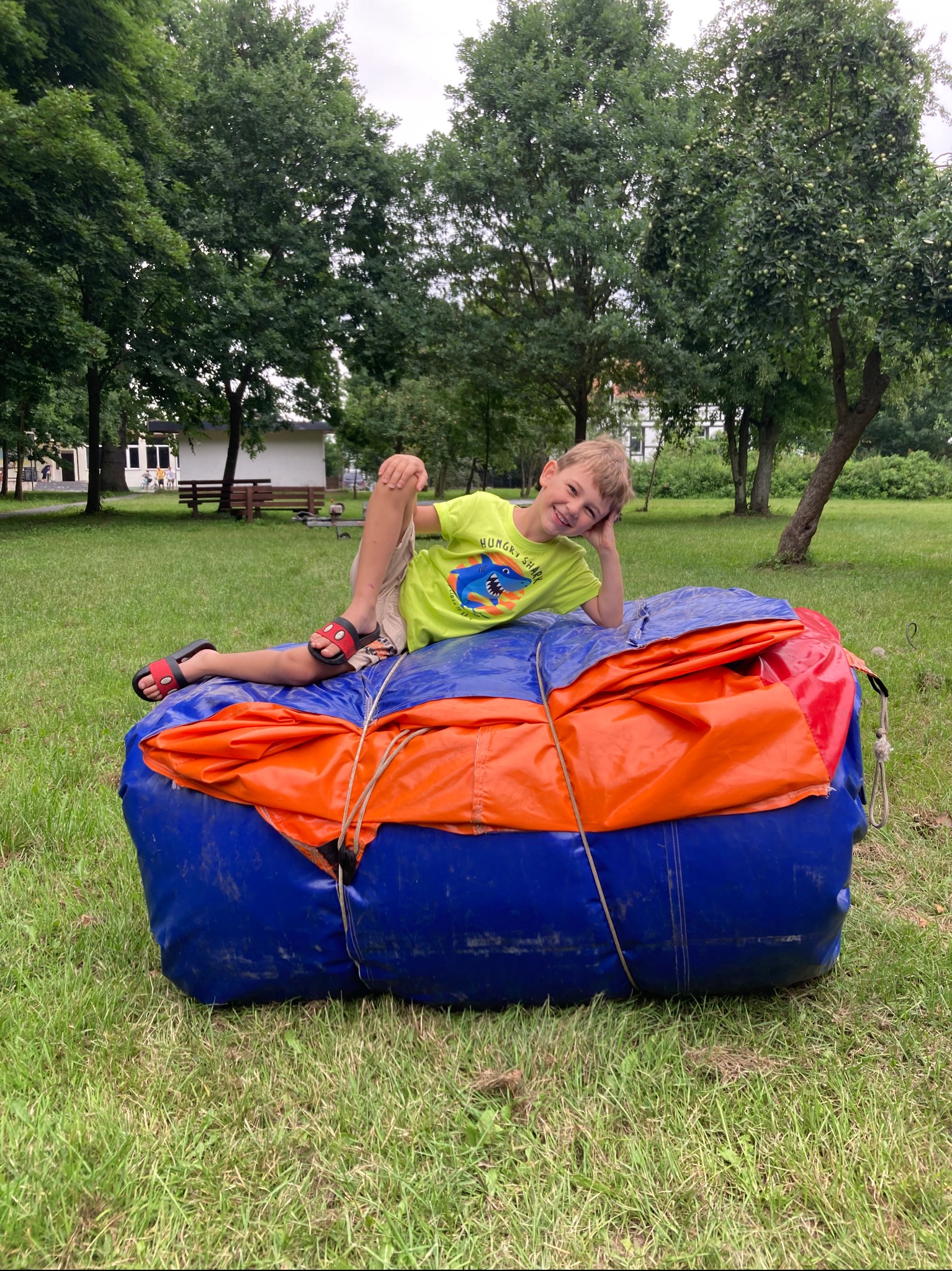 Uśmiechnięty chłopiec w zielonej koszulce z rekinem leży na zwiniętym, niebiesko-pomarańczowym dmuchanym zamku na trawie w parku.