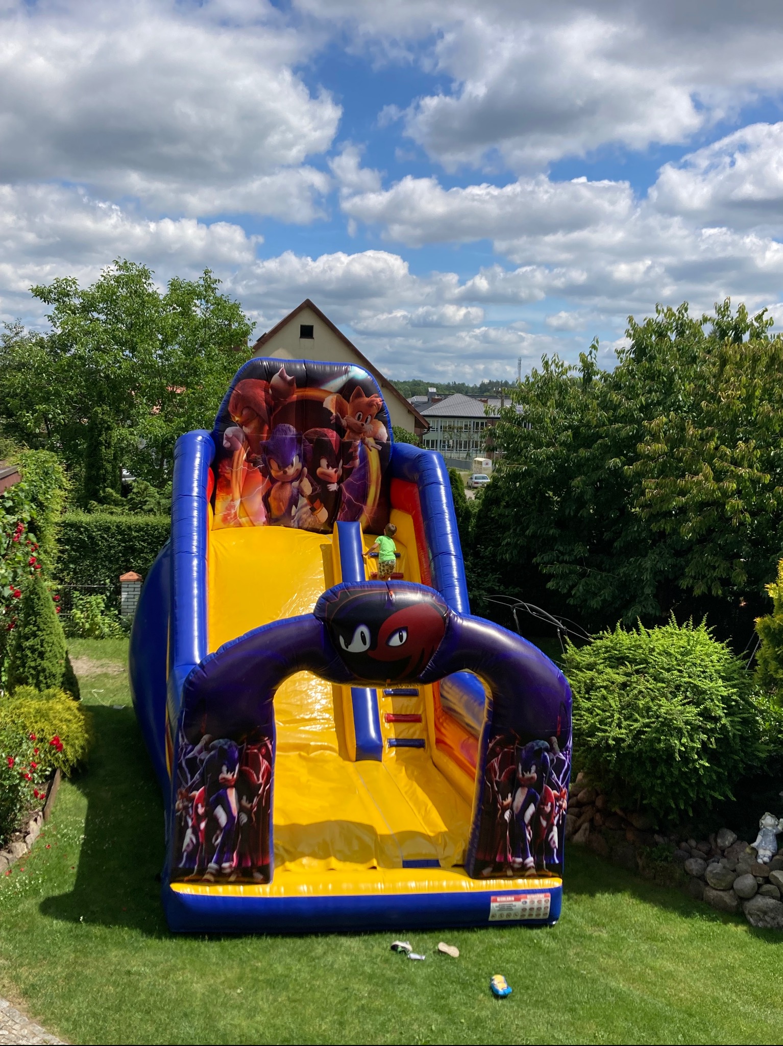Niebiesko-żółta dmuchana zjeżdżalnia z motywem postaci z gier wideo ustawiona na trawniku w ogrodzie, widoczna osoba na górze zjeżdżalni