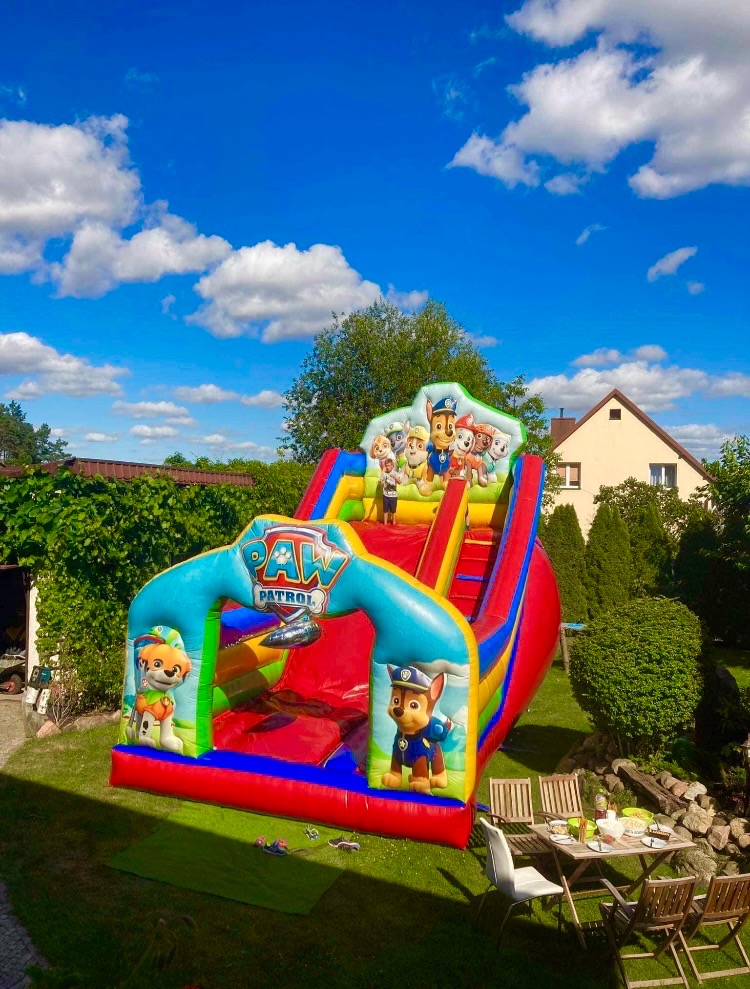 Kolorowa dmuchana zjeżdżalnia z motywem bajki dla dzieci, ustawiona na trawniku w ogrodzie, w tle błękitne niebo z białymi chmurami i dom.