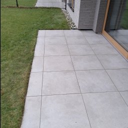 A long way - Ułożony taras z jasnoszarych, kwadratowych płyt betonowych przylegający do budynku z szarej cegły, widok z góry na trawnik i ogrodzenie.