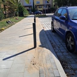 A long way - Świeżo ułożona, szara kostka brukowa wzdłuż ulicy z zamontowanymi czarnymi słupkami odgradzającymi; widoczny piasek wokół słupków i na powierzchni kostki; w tle budynki mieszkalne i zaparkowany...