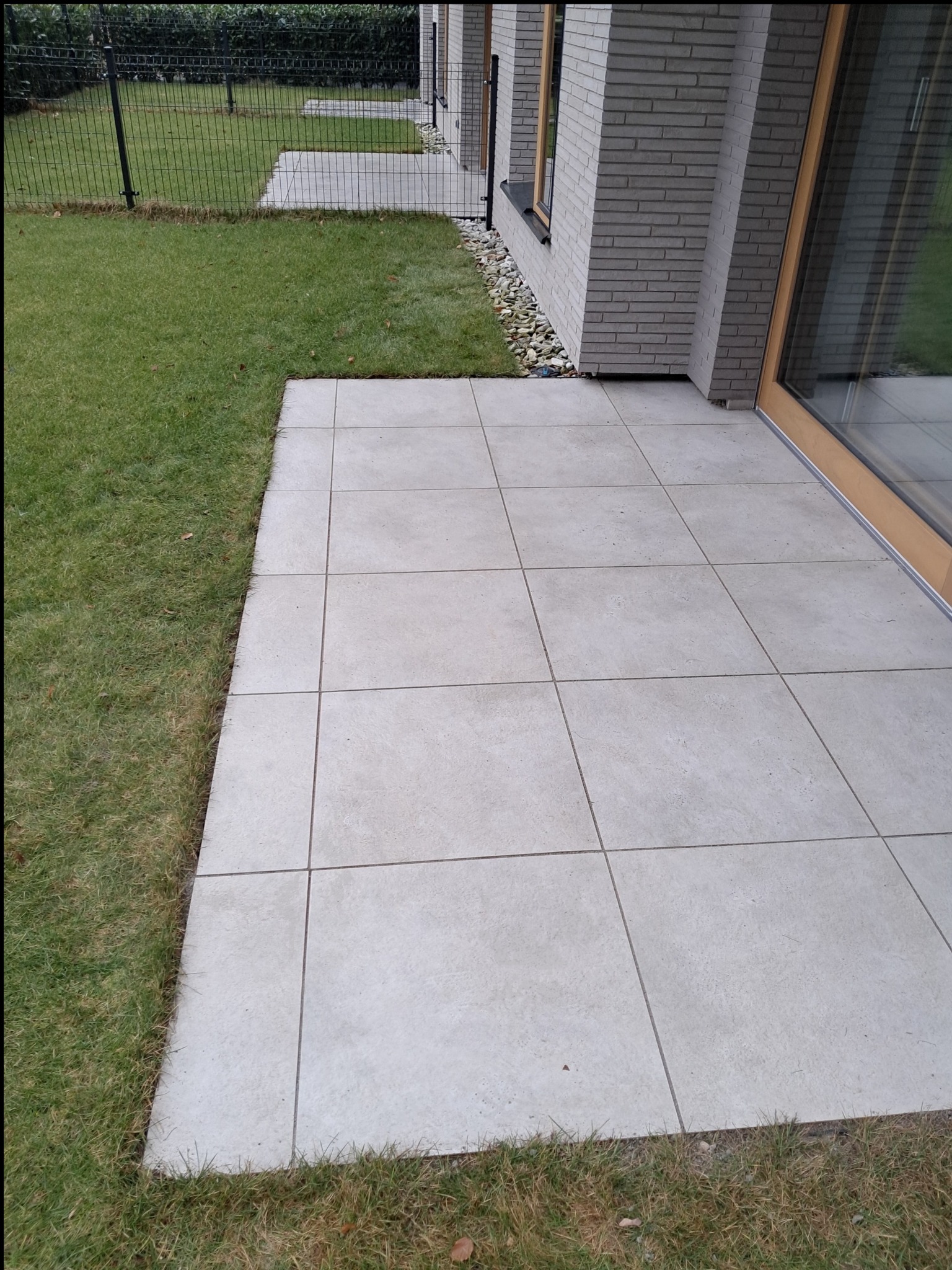 Ułożony taras z jasnoszarych, kwadratowych płyt betonowych przylegający do budynku z szarej cegły, widok z góry na trawnik i ogrodzenie.