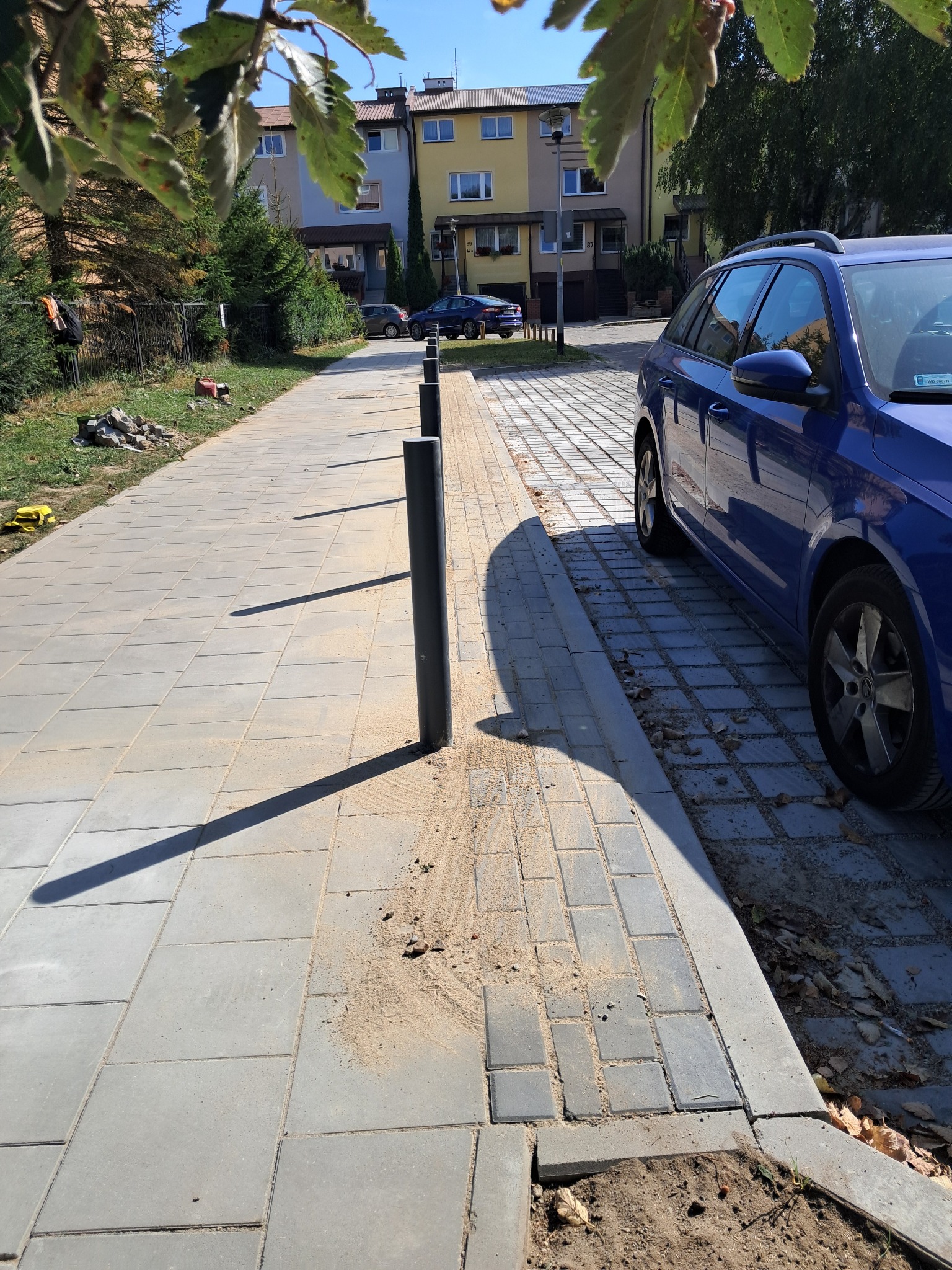 Świeżo ułożona, szara kostka brukowa wzdłuż ulicy z zamontowanymi czarnymi słupkami odgradzającymi; widoczny piasek wokół słupków i na powierzchni kostki; w tle budynki mieszkalne i zaparkowany...