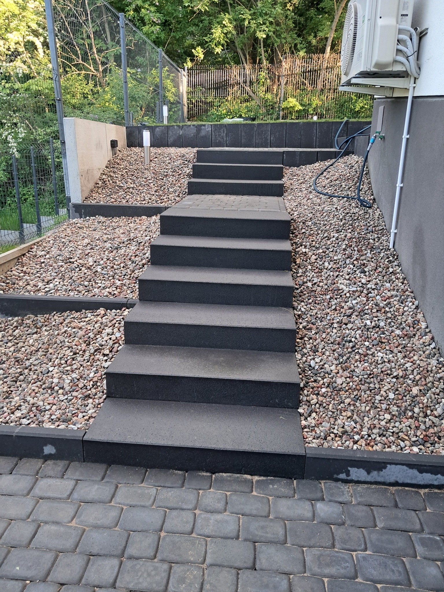 Schody z prefabrykatów betonowych prowadzące w górę, otoczone kamieniami ozdobnymi i kostką brukową. Widoczny fragment budynku i ogrodzenie.