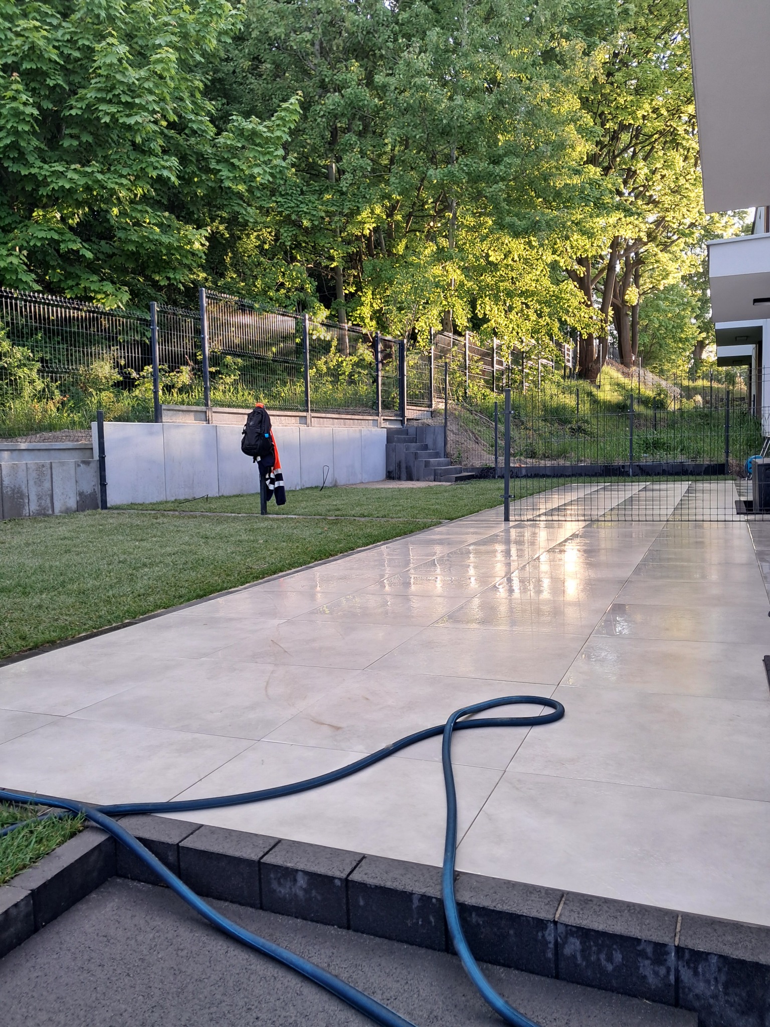 Jasne płytki chodnikowe, mokre po umyciu, z niebieskim wężem ogrodowym w rogu. W tle trawnik, ogrodzenie i drzewa.