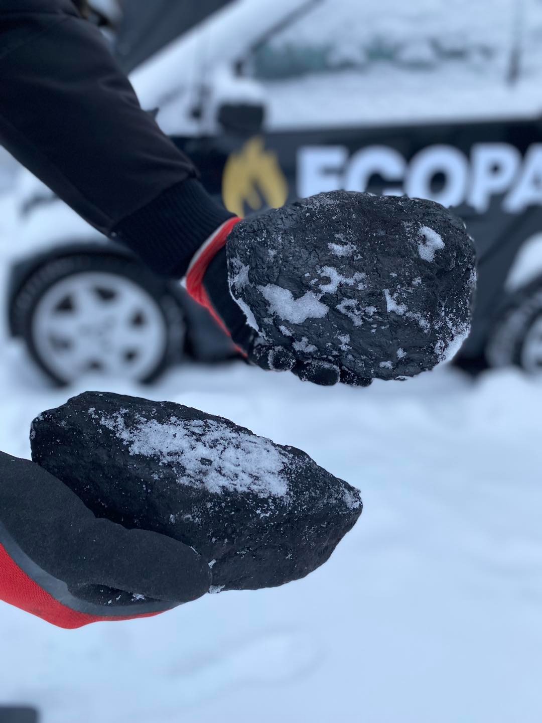 Dwie bryły czarnego węgla z białym śniegiem w rękawicach na tle zasypanego śniegiem samochodu z logo ECOPA.