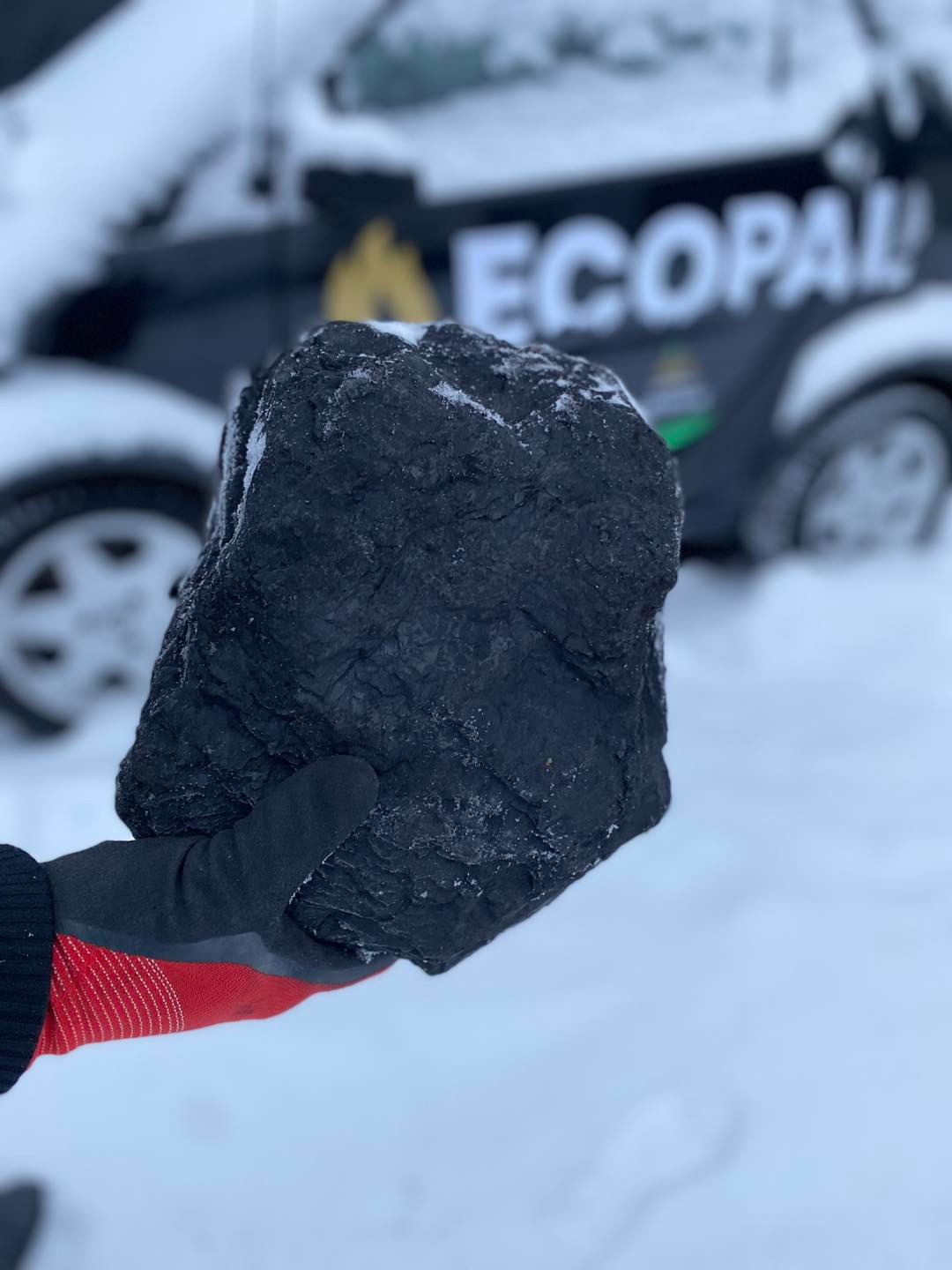 Bryła czarnego węgla, pokryta śniegiem, trzymana w dłoni w rękawicy roboczej z czerwonymi elementami, na tle ośnieżonego terenu i samochodu z logo 'ECOPAL'.