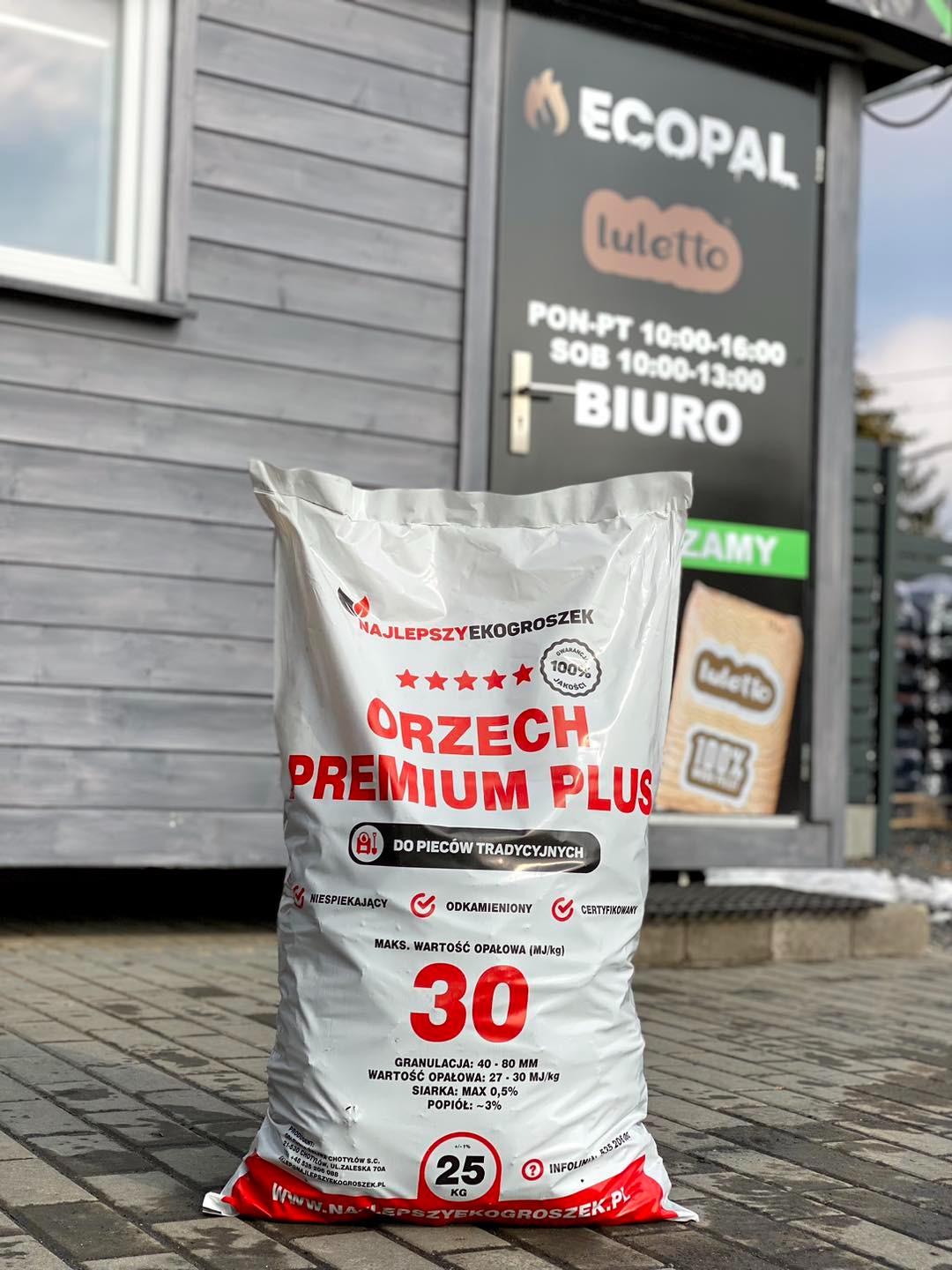 Worek z ekogroszkiem 'Orzech Premium Plus' 25 kg, z widocznymi informacjami o granulacji, wartości opałowej i zawartości siarki, ustawiony przed budynkiem biura firmy 'Ecopal' z godzinami otwarcia.