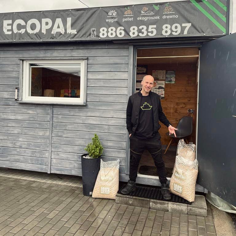 Przedsiębiorca prezentuje dwa worki pelletu przed wejściem do siedziby firmy 'ECOPAL', oferującej również węgiel, ekogroszek i drewno kominkowe; widoczna tablica reklamowa z numerem telefonu.