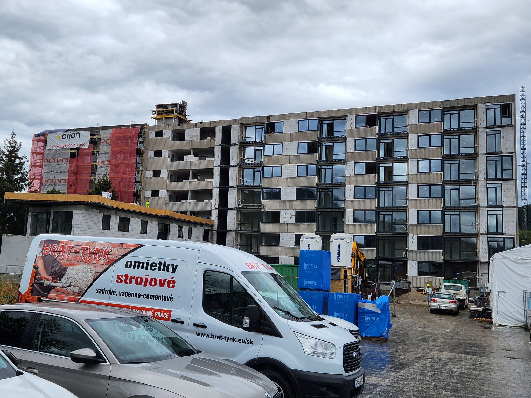 Budynek w trakcie budowy z widocznym rusztowaniem i reklamą dewelopera, na pierwszym planie samochody firmowe z logo firmy oferującej usługi tynkarskie.