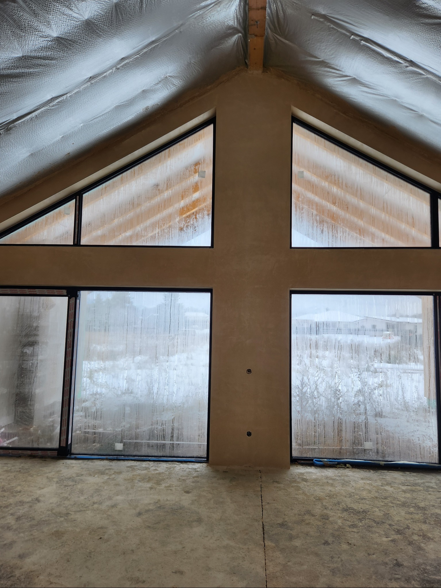 Wnętrze domu w trakcie prac wykończeniowych: sufit ocieplony folią, ściany pokryte świeżym jasnym tynkiem, duże okna z widokiem na zewnątrz pokryte parą wodną.