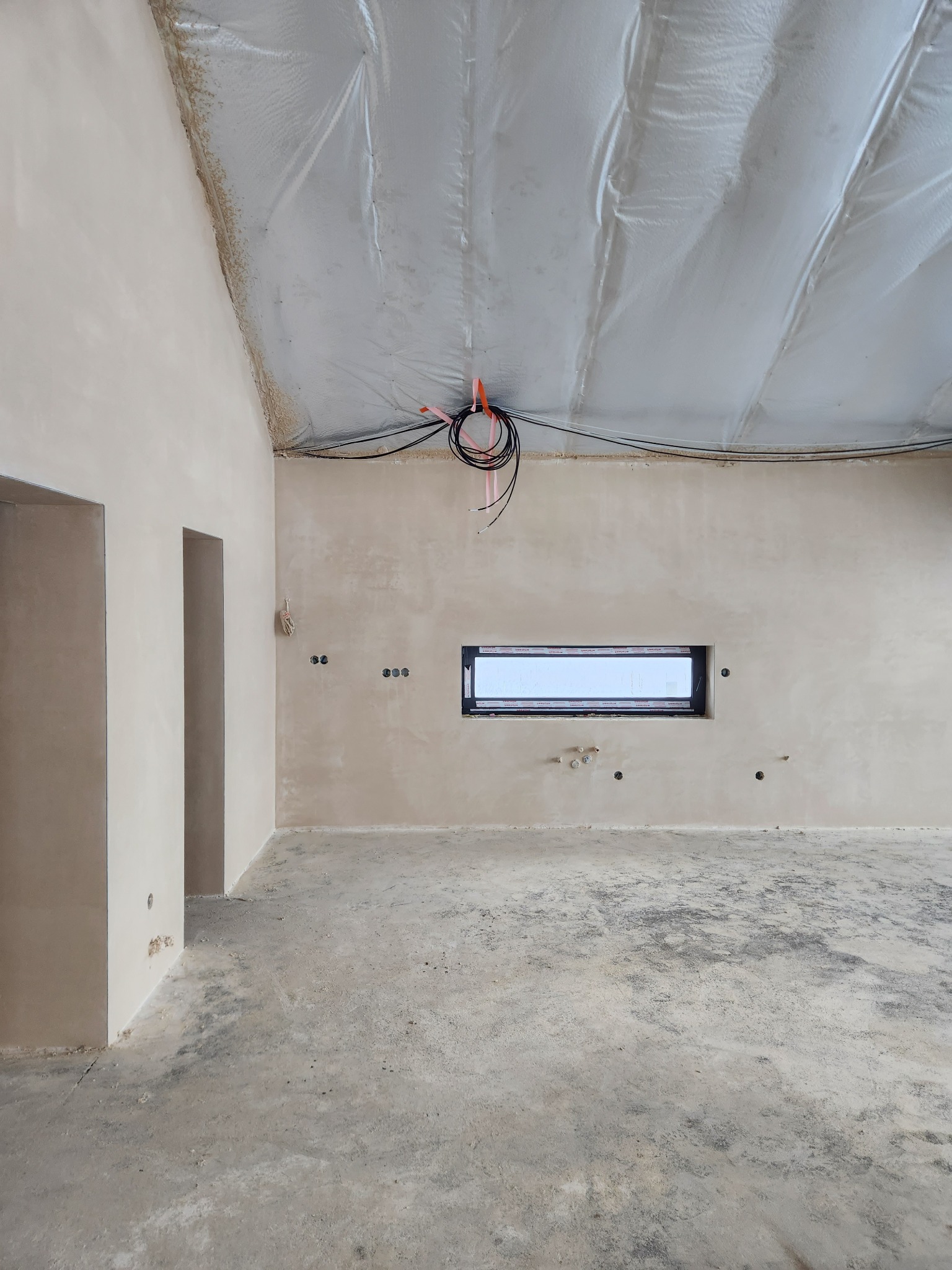 Wnętrze surowego domu po tynkach maszynowych, widoczne ściany z gładzią, okno, instalacja elektryczna pod sufitem i betonowa podłoga.