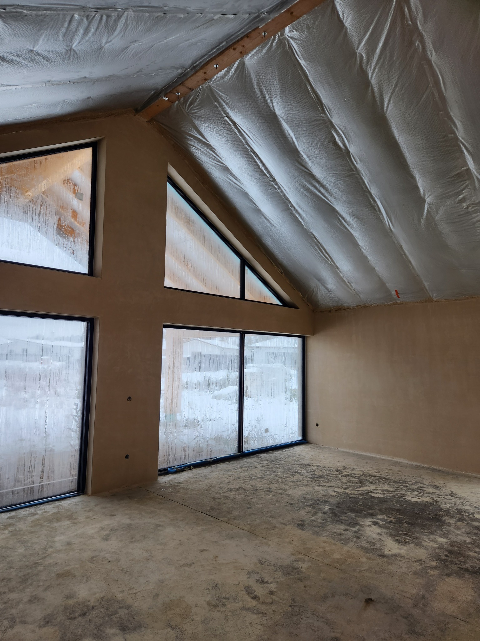 Wnętrze domu w stanie surowym zamkniętym po nałożeniu tynków, widoczne okna z czarnymi ramami, ściany otynkowane na beżowo, sufit pokryty folią termoizolacyjną, podłoga betonowa. Na szybach...