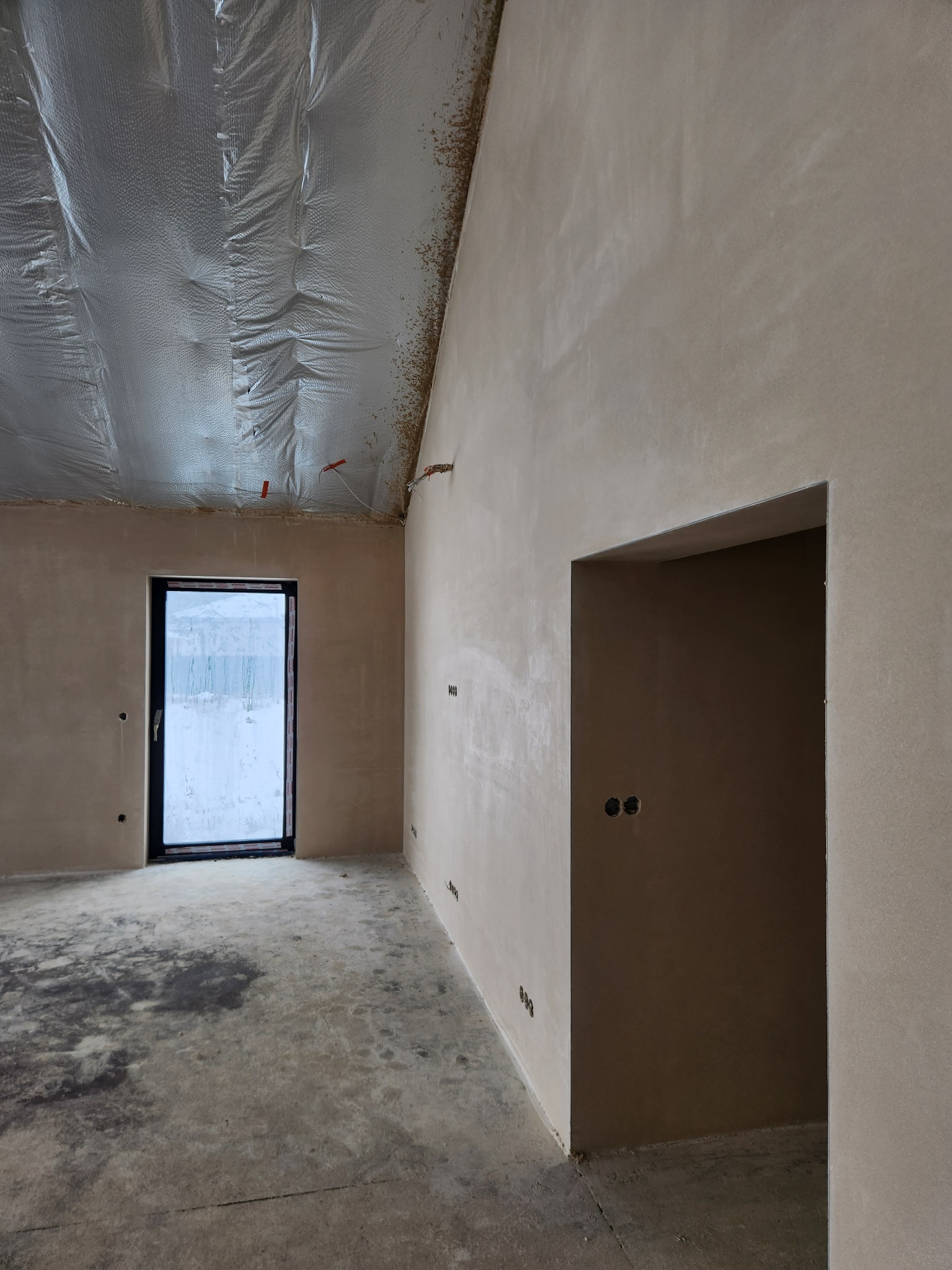 Wnętrze surowego domu po nałożeniu gładzi gipsowej na ściany, widoczne puszki elektryczne i folia izolacyjna na suficie, okno z widokiem na zaśnieżony ogród.