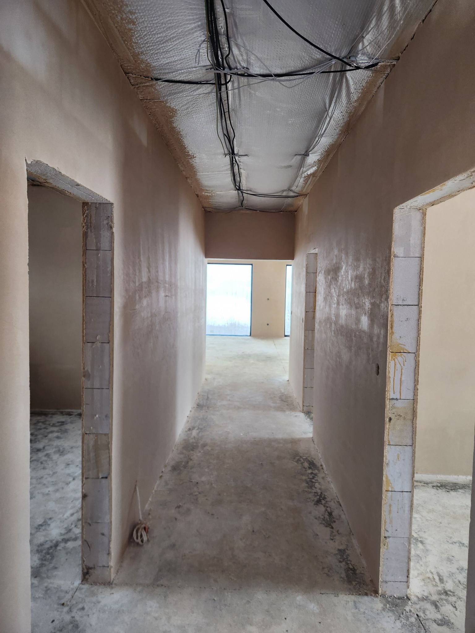 Wnętrze w trakcie prac wykończeniowych: świeżo otynkowane ściany w odcieniach beżu, sufit z widoczną izolacją i przewodami elektrycznymi, surowa betonowa podłoga oraz otwory drzwiowe z widocznymi...