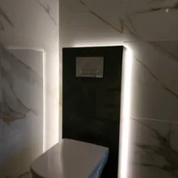 Nowoczesna toaleta podwieszana z czarnym panelem spłukującym, otoczona białymi płytkami marmurkowymi i podświetleniem LED.