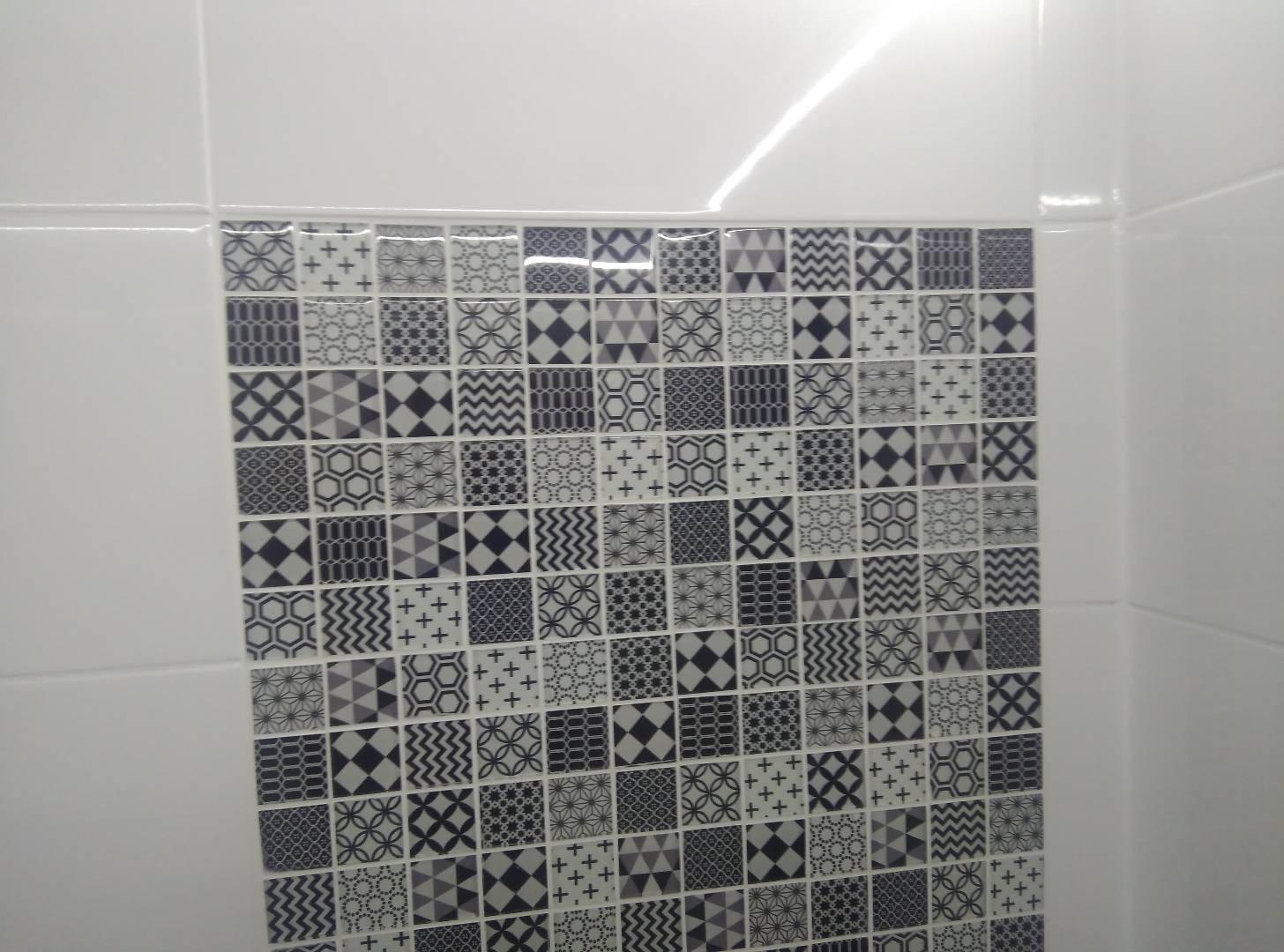 Fragment ściany łazienki wyłożony białymi płytkami ceramicznymi z dekoracyjnym pasem mozaiki w odcieniach szarości z geometrycznymi wzorami.