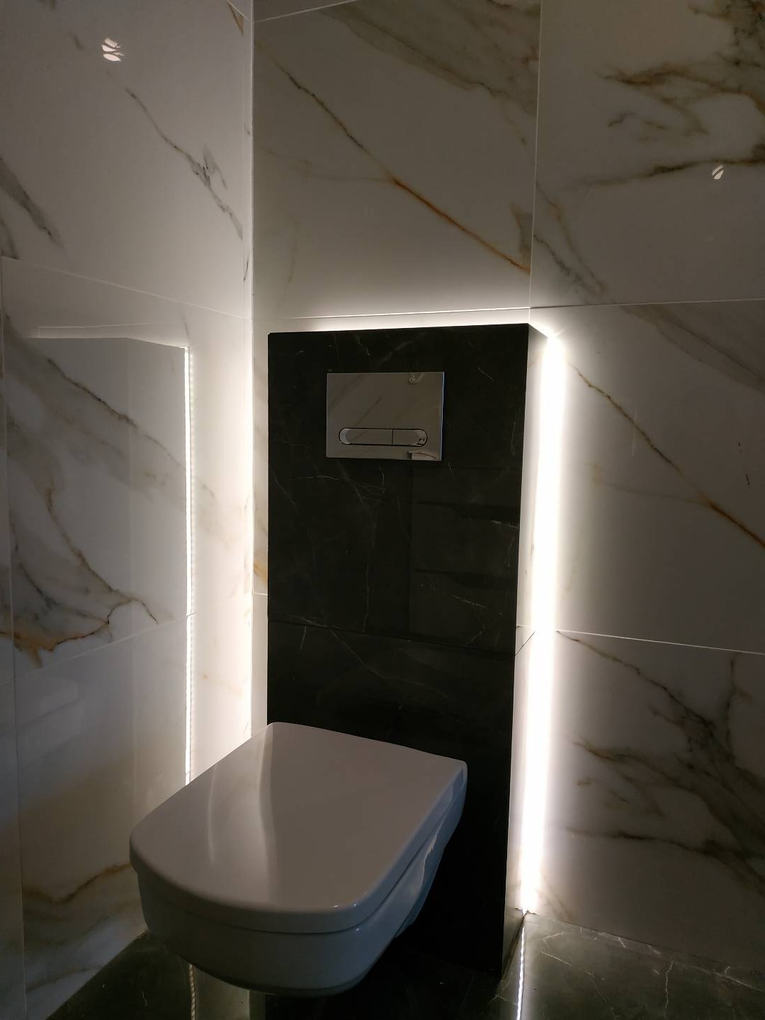 Nowoczesna toaleta podwieszana z czarnym panelem spłukującym, otoczona białymi płytkami marmurkowymi i podświetleniem LED.