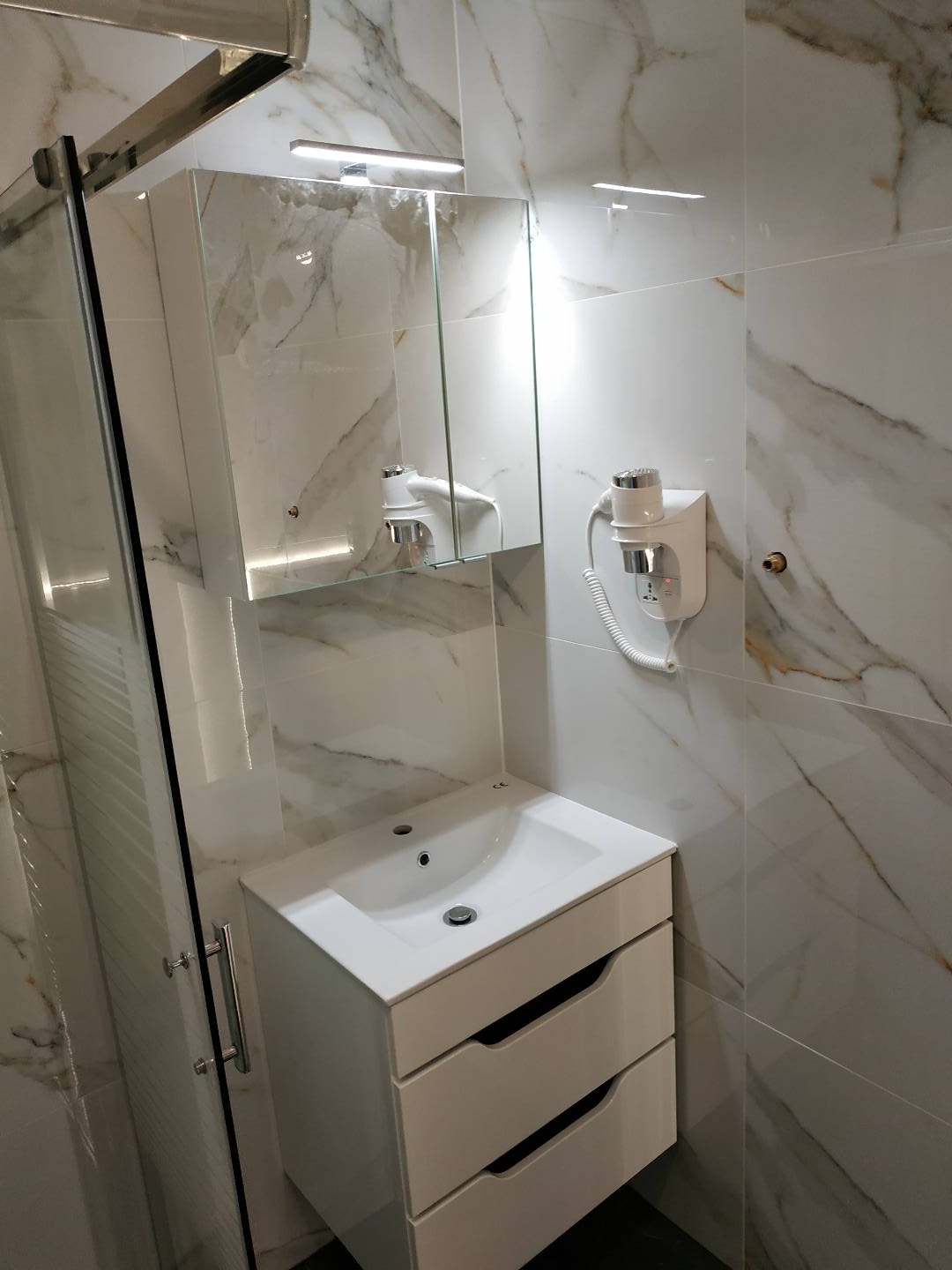 Biała umywalka z szafką z trzema szufladami w łazience wykończonej płytkami imitującymi marmur, obok kabina prysznicowa i suszarka do włosów na ścianie.