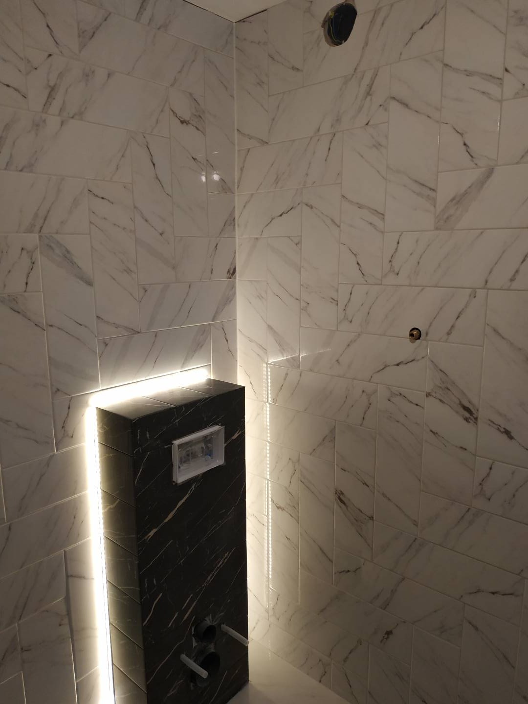 Ściana łazienki wyłożona białymi płytkami imitującymi marmur, z czarną obudową stelaża podtynkowego WC, podświetloną pionowymi paskami LED po bokach.