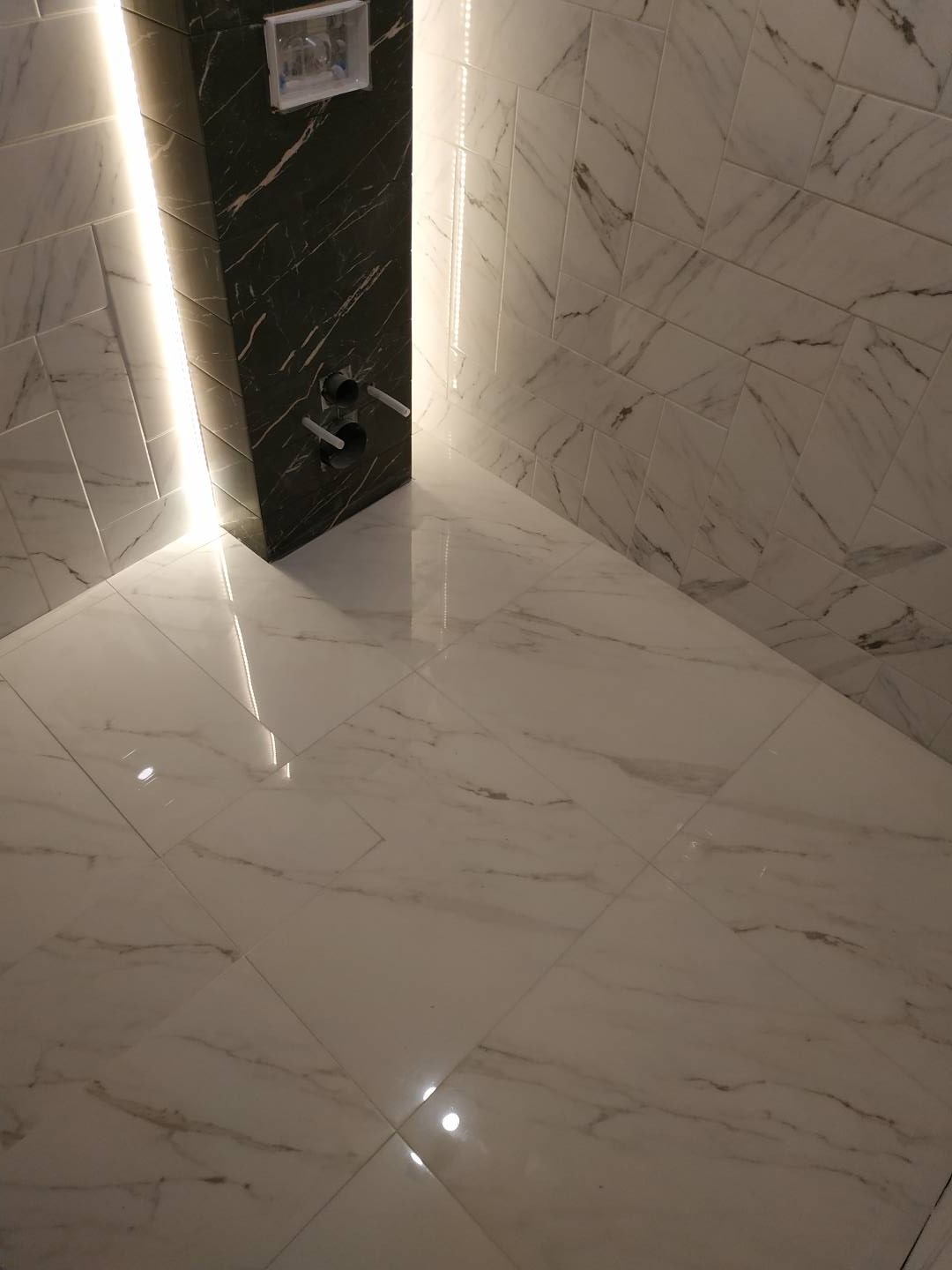 Nowoczesna łazienka z białymi płytkami imitującymi marmur, czarnym elementem zabudowy geberitu z podświetleniem LED i widocznymi rurami doprowadzającymi wodę.