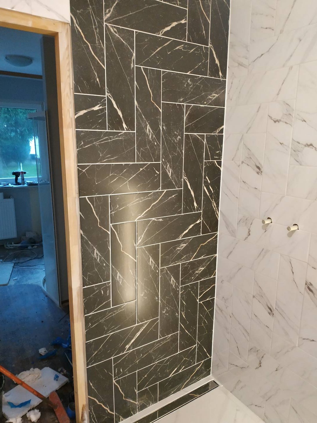 Częściowo wykończona łazienka z czarnymi i białymi marmurkowymi płytkami ułożonymi w różne wzory; widoczne niedokończone obramowanie drzwi i elementy instalacji.