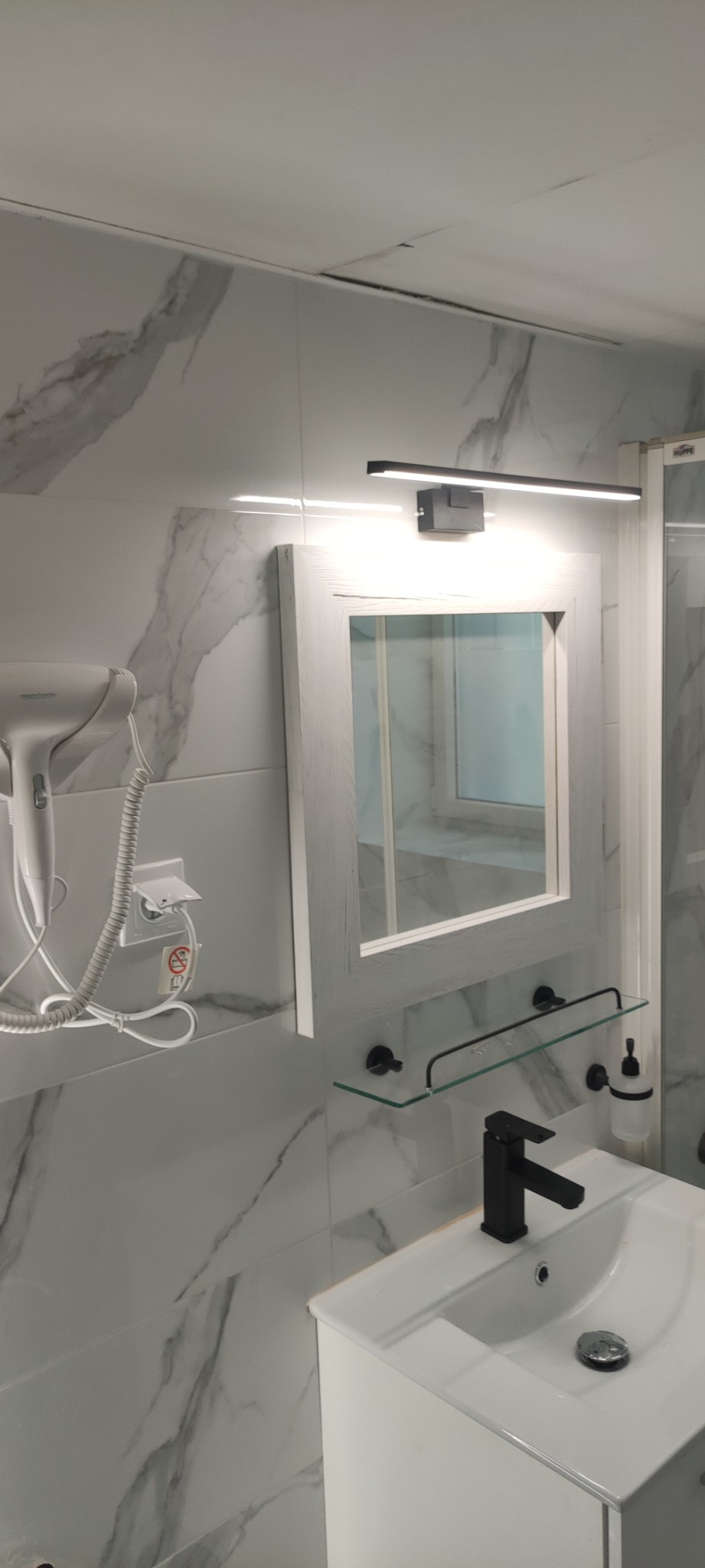 Nowoczesna łazienka z marmurowymi płytkami, czarną armaturą i podświetlanym lustrem, suszarka do włosów zamontowana na ścianie.