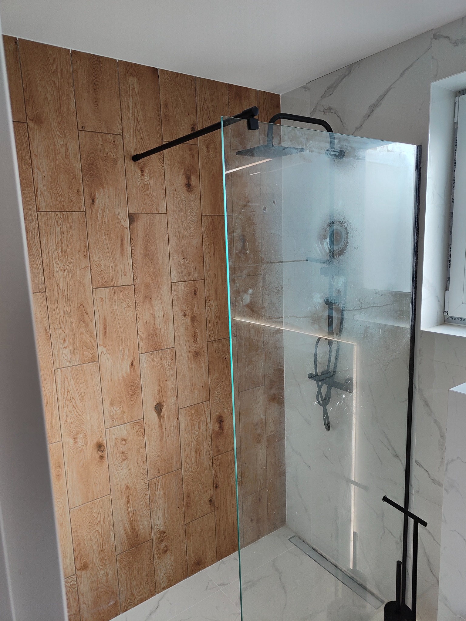 Nowoczesna kabina prysznicowa z czarną armaturą i ścianą wyłożoną płytkami imitującymi drewno, widoczny odpływ liniowy i marmurowe płytki na pozostałych ścianach.