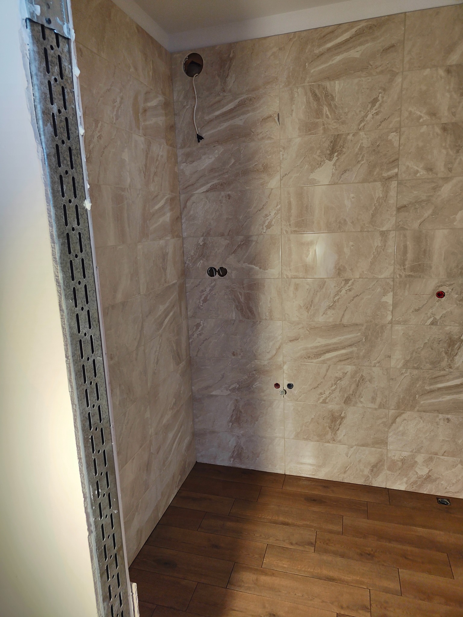 Wnętrze łazienki w Kołobrzegu w trakcie prac wykończeniowych: ułożone płytki imitujące marmur na ścianach, drewnopodobne panele na podłodze, widoczne puszki elektryczne i metalowa konstrukcja przy...