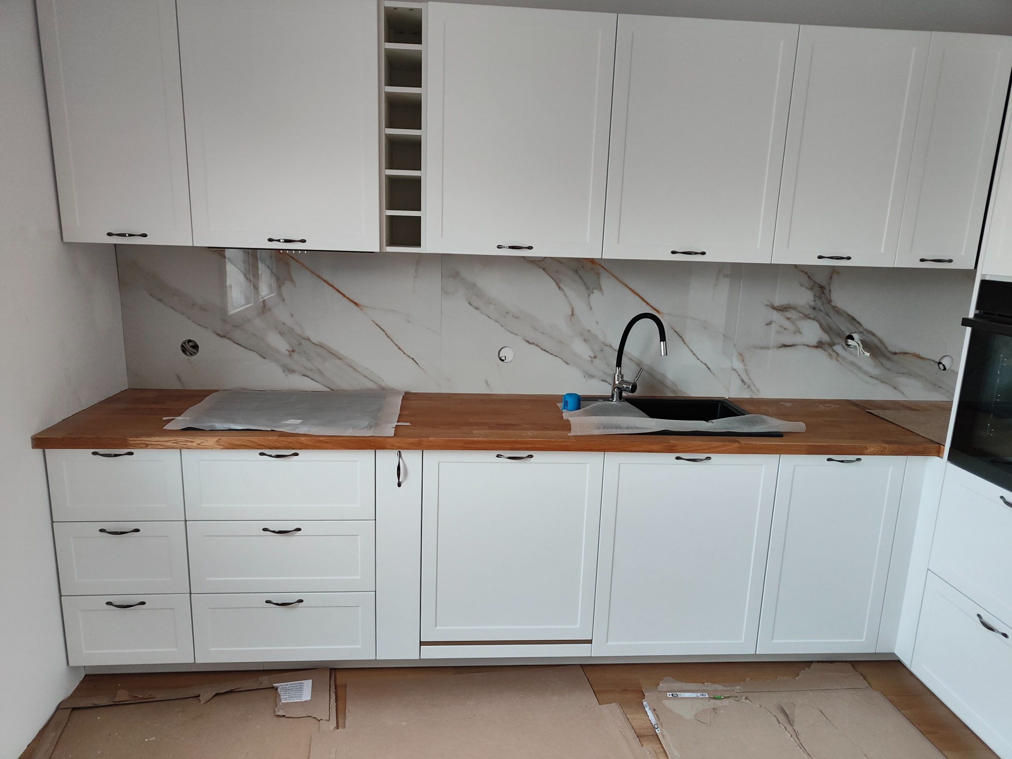 Biała zabudowa kuchenna z drewnianym blatem i marmurowym backsplashiem, widoczne przygotowania do podłączenia armatury, podłoga zabezpieczona kartonem.