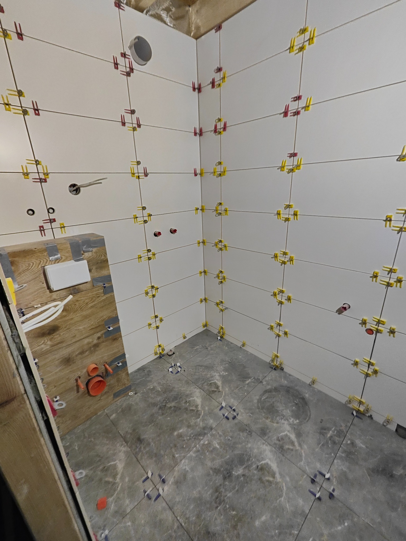 Świeżo wyłożona, biała glazura ścienna w łazience, z widocznymi żółtymi i czerwonymi klipsami poziomującymi. Na podłodze szare płytki imitujące marmur, również z klipsami. Widoczna instalacja...