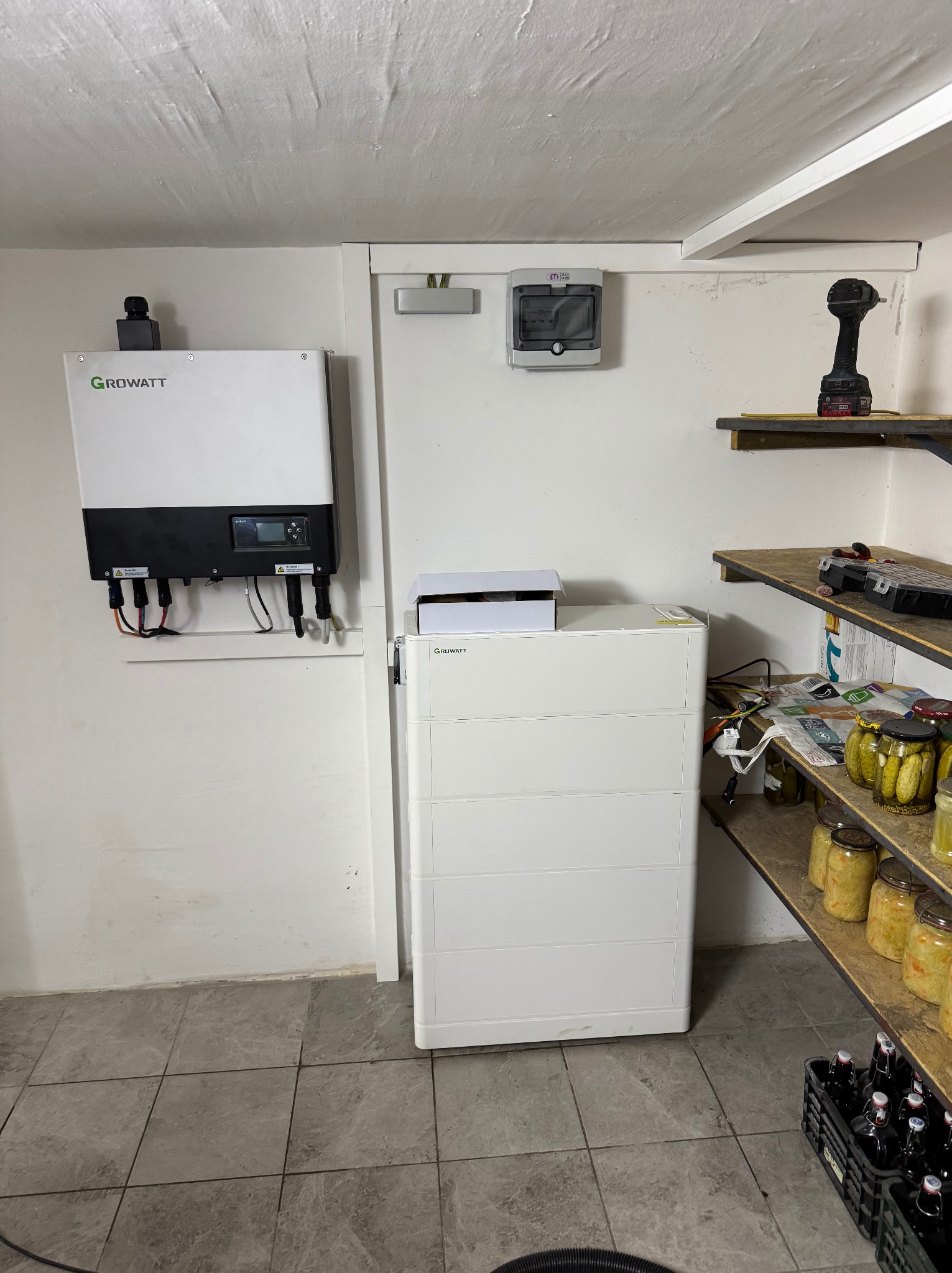 Zamontowany inwerter Growatt na białej ścianie piwnicy obok białej baterii energii, na półkach słoiki z przetworami i skrzynka z butelkami piwa.