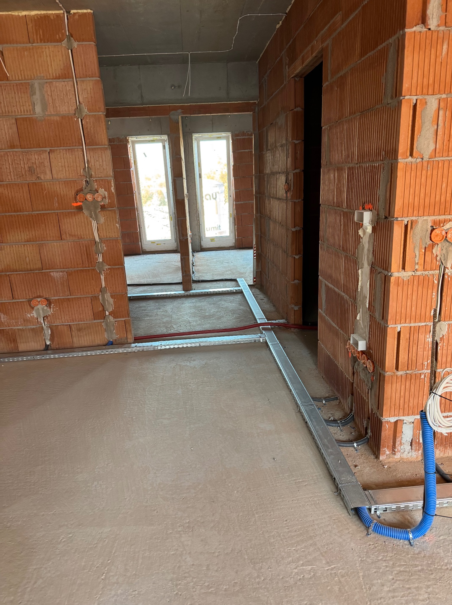 Surowy stan wnętrza budynku w trakcie prac instalacyjnych: widoczne przewody elektryczne w korytach podłogowych i puszkach na ścianach z cegły dziurawki, świeżo wylana wylewka podłogowa.