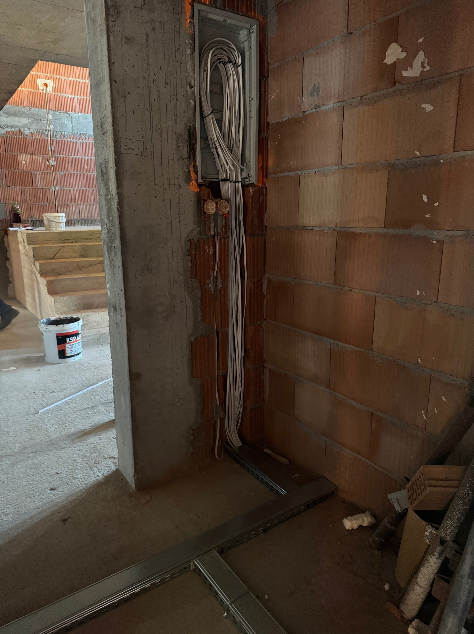 Wiązka białych kabli elektrycznych wychodząca z metalowej puszki w surowej ścianie z czerwonej cegły, widoczne przewody prowadzone wzdłuż ściany, w tle betonowy słup i schody w stanie surowym.