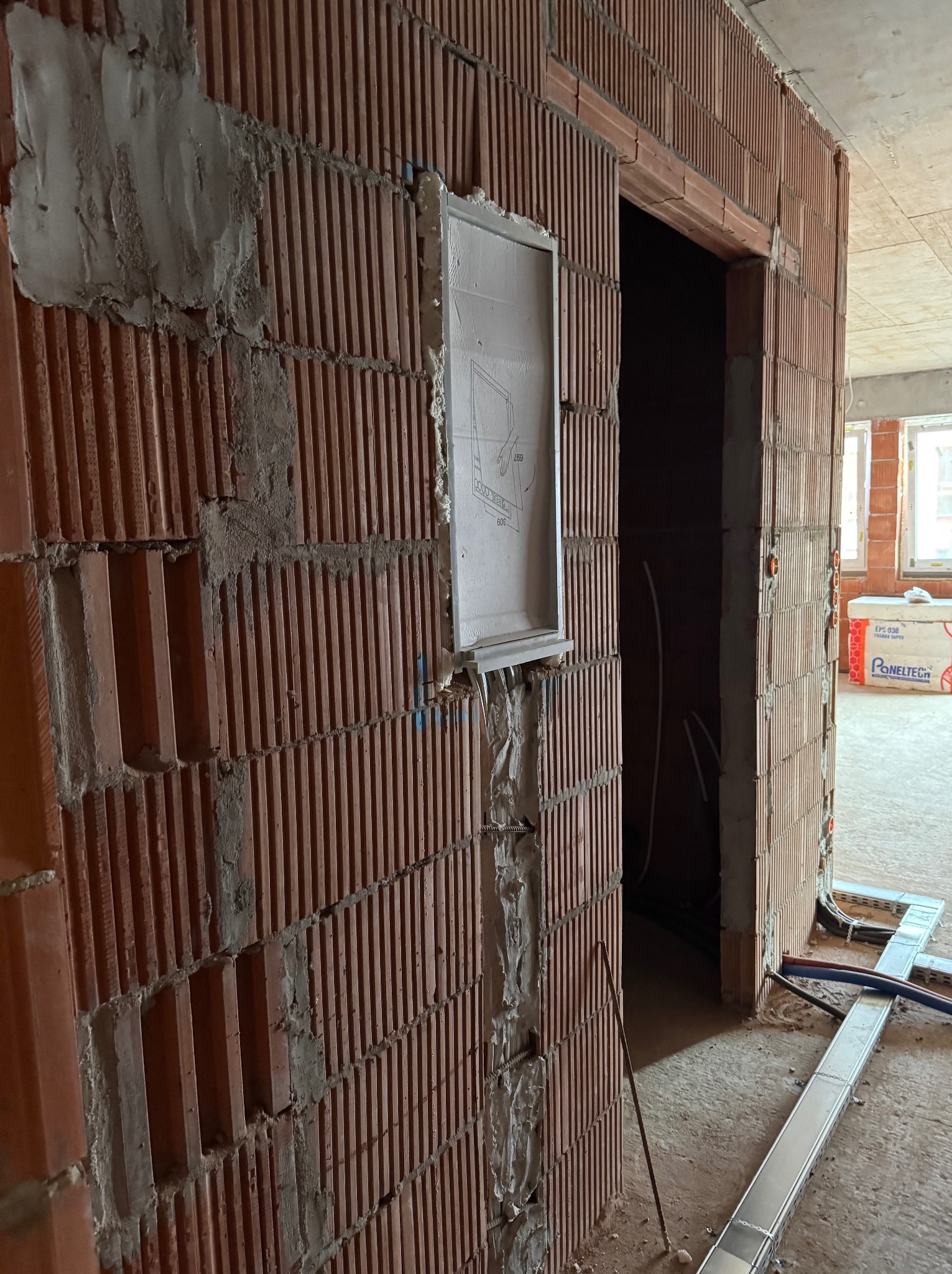 Montaż skrzynki elektrycznej podtynkowej w ścianie z cegły szczelinowej, widoczne przewody i koryto kablowe na podłodze w surowym stanie.