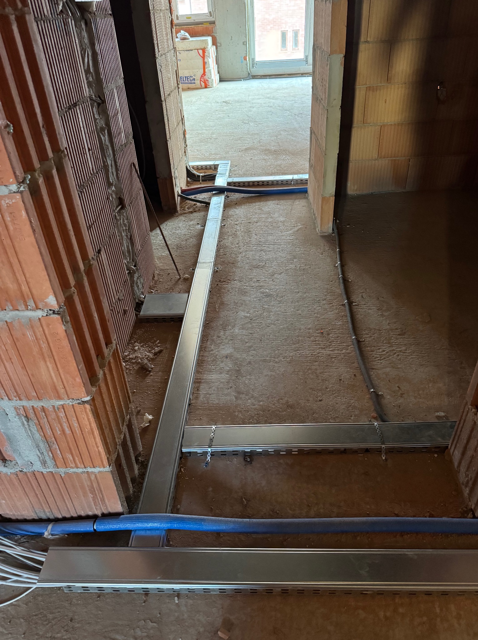 Wnętrze w trakcie prac wykończeniowych: prowadzenie kabli w metalowych korytkach pod podłogą, widoczne ceglane ściany i otwór drzwiowy.