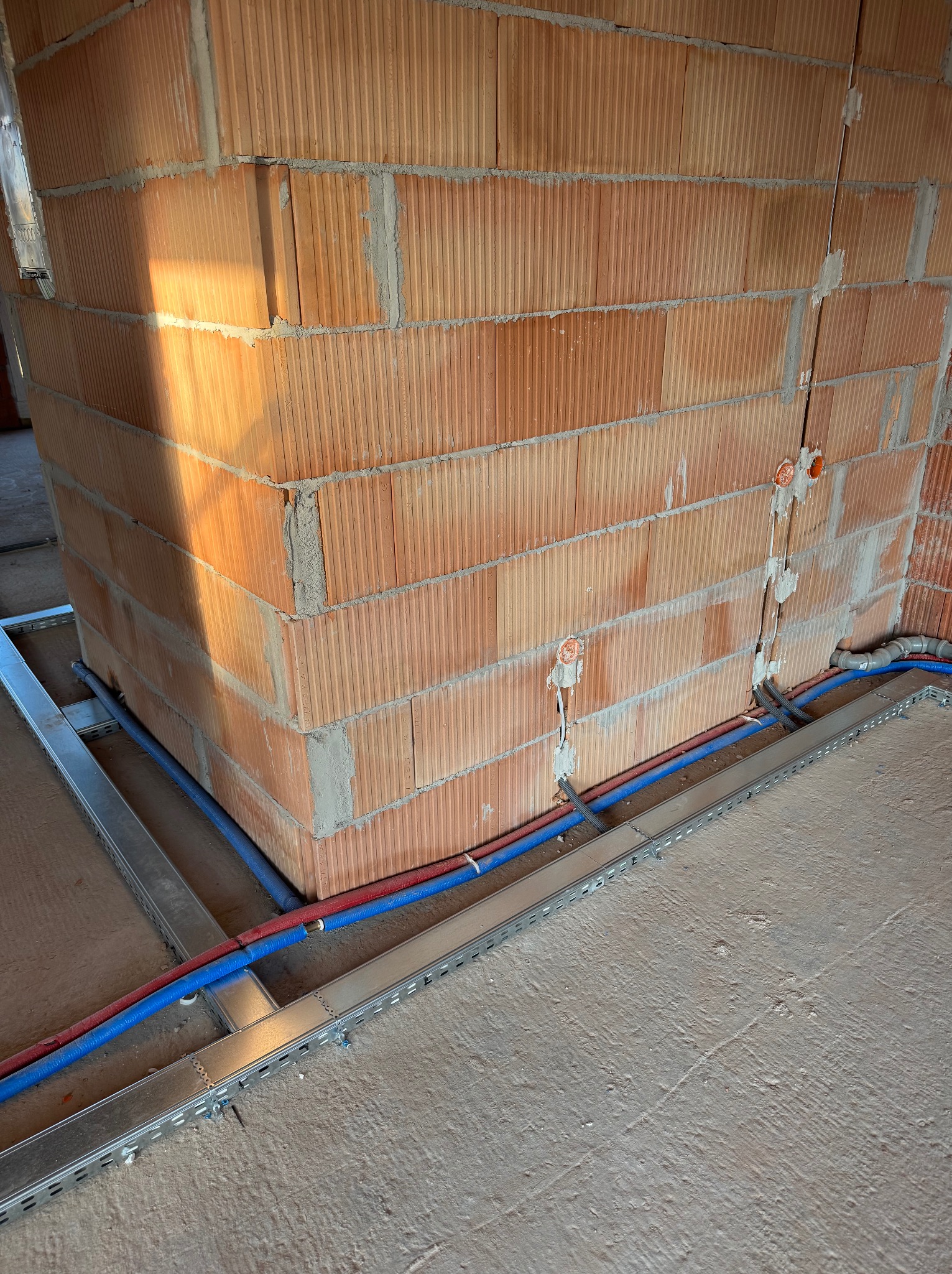 Czerwona cegła, instalacja rur w korytkach podłogowych, widoczne puszki elektryczne w ścianie, stan surowy.