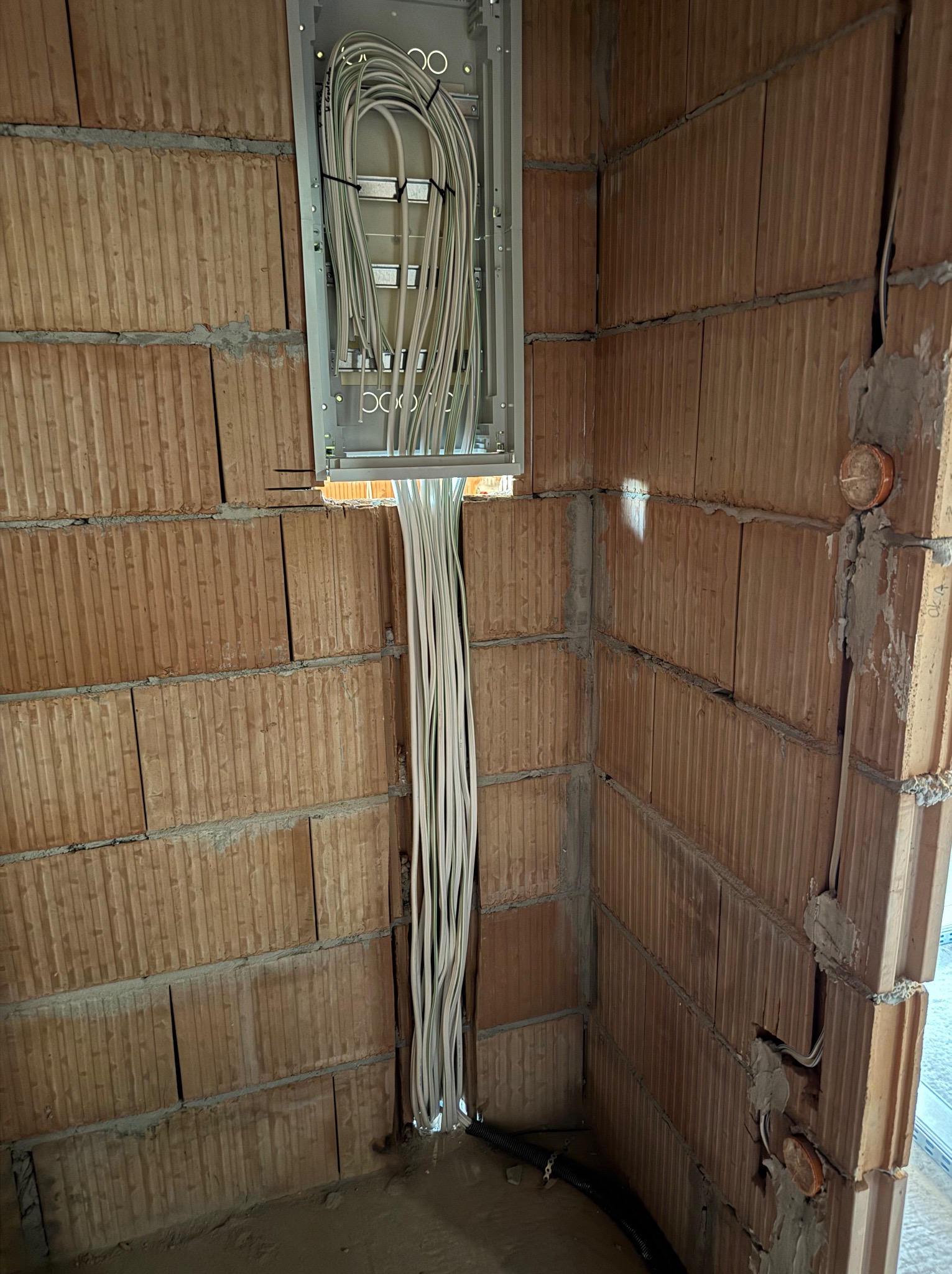 Rozdzielnica elektryczna z wyprowadzonymi przewodami w surowym wnętrzu z cegły dziurawki, widoczne mocowania kabli i fragment rury instalacyjnej w podłodze.